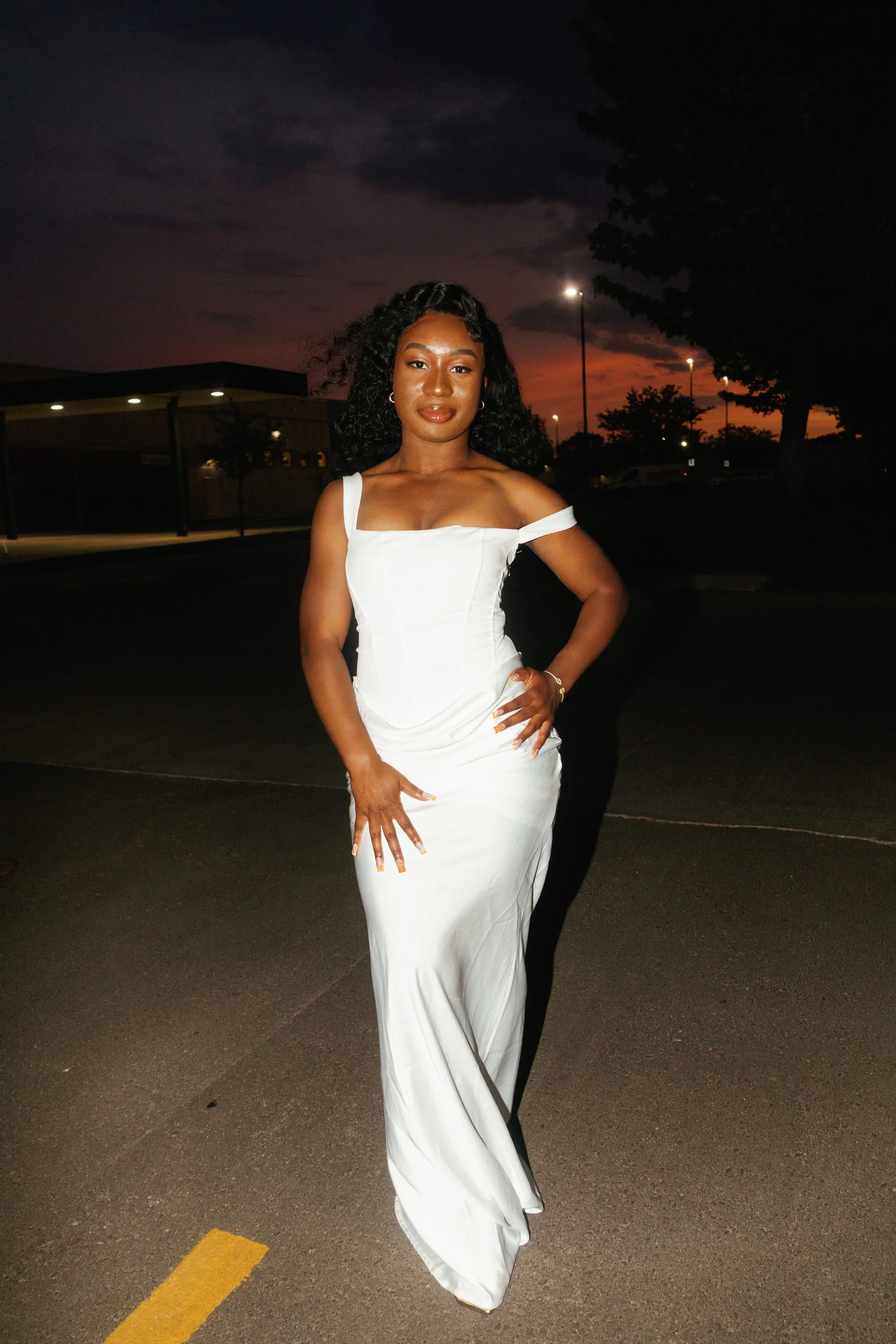 A woman in a white dress standing outdoors during sunset with trees and parking lot in the background.