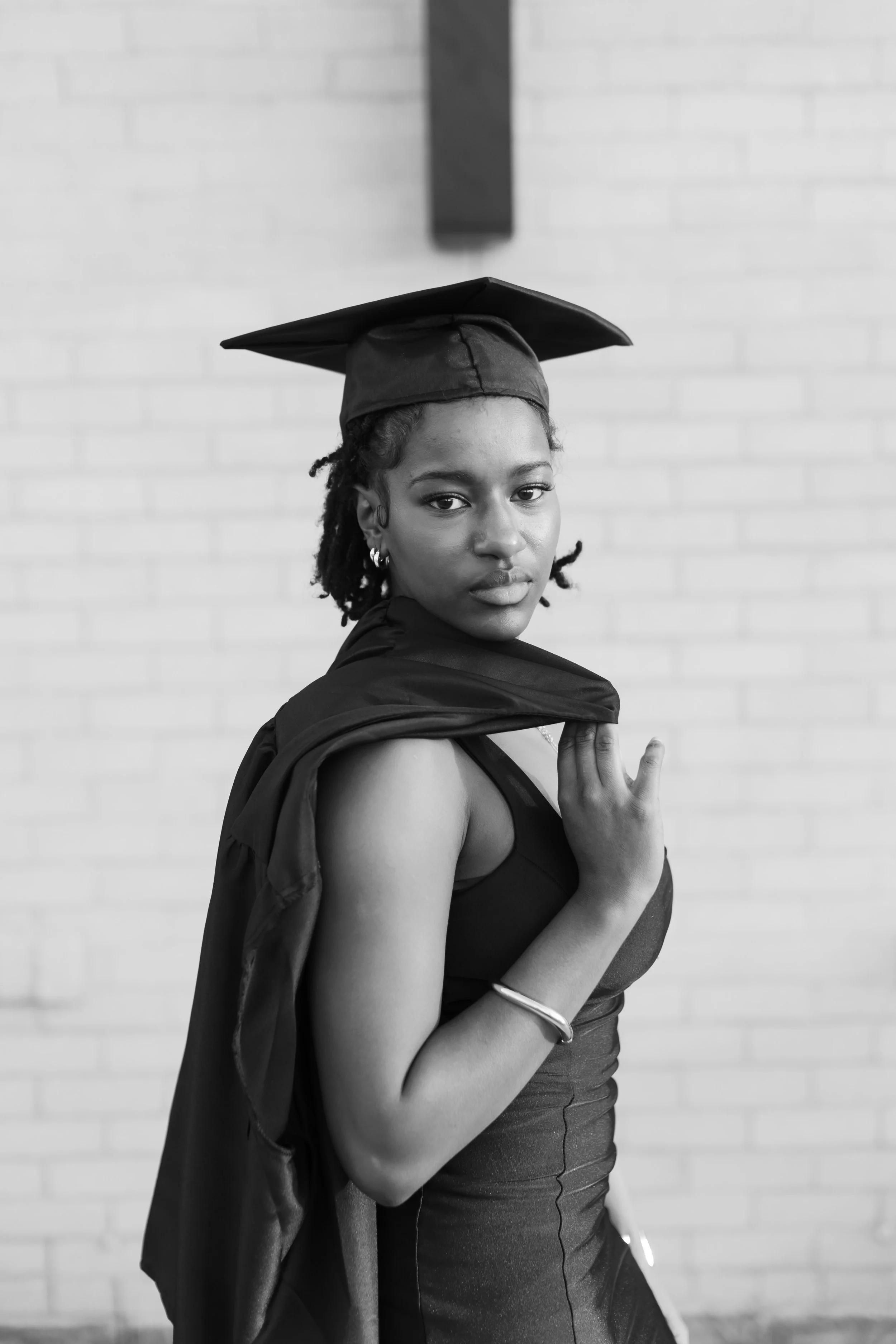 A young woman wearing a graduation cap and gown, holding a diploma, standing in front of a white brick wall.
