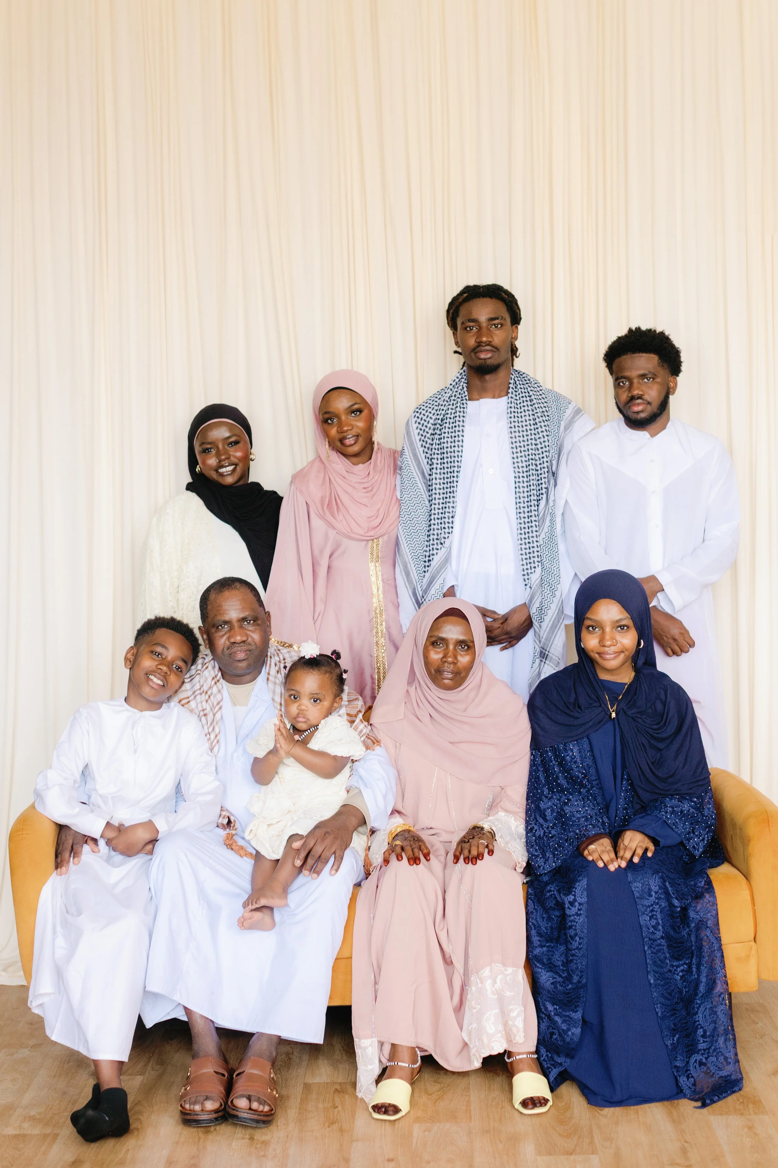 A group of people, including adults and children, posing together for a photo in front of a cream-colored curtain. They are dressed in traditional African attire, with some seated on a yellow couch and others standing behind.