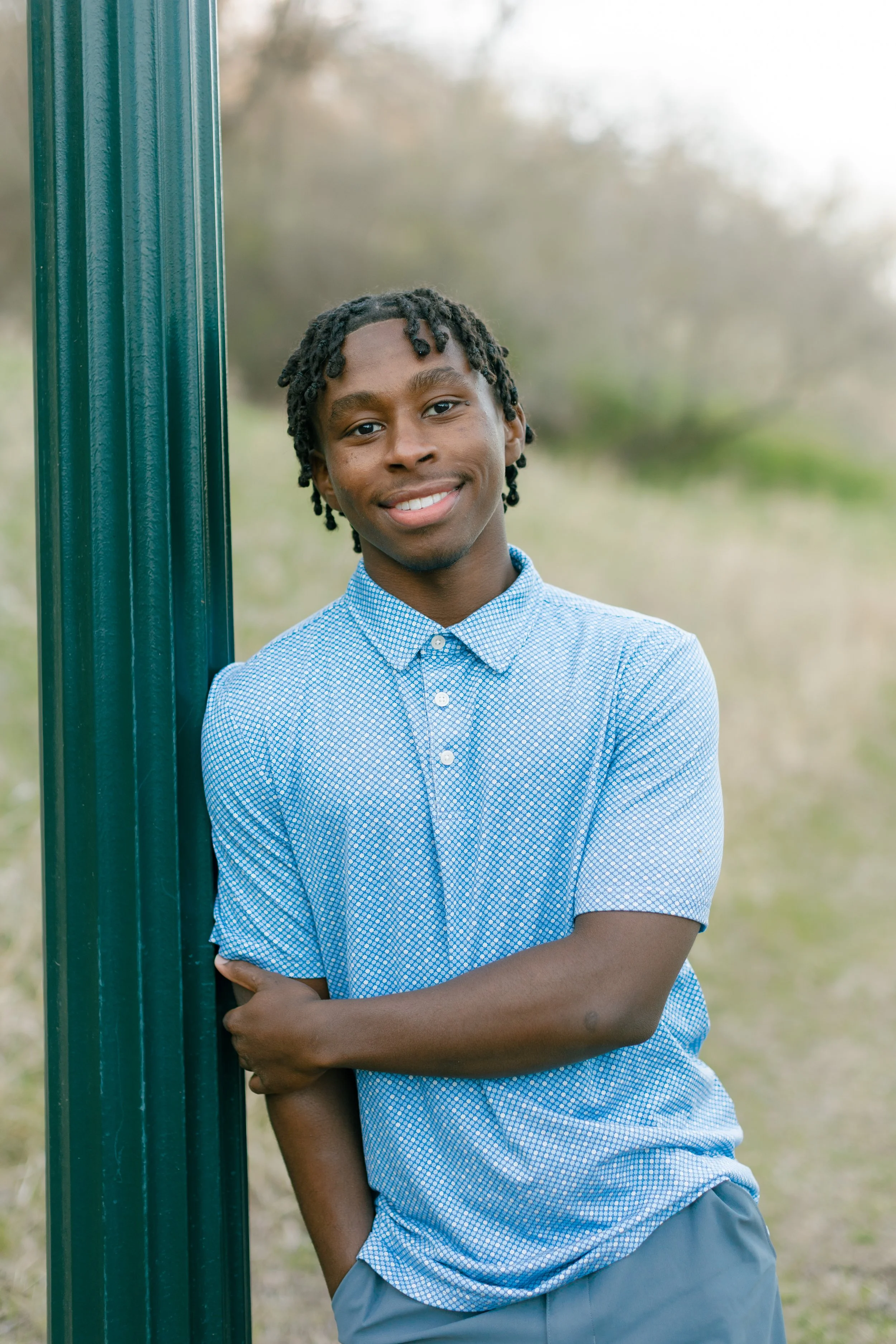 A young man with earrings and dreadlocks leaning against a green metal pole outdoors, smiling at the camera, wearing a blue checkered shirt and gray shorts, with a blurred natural background.