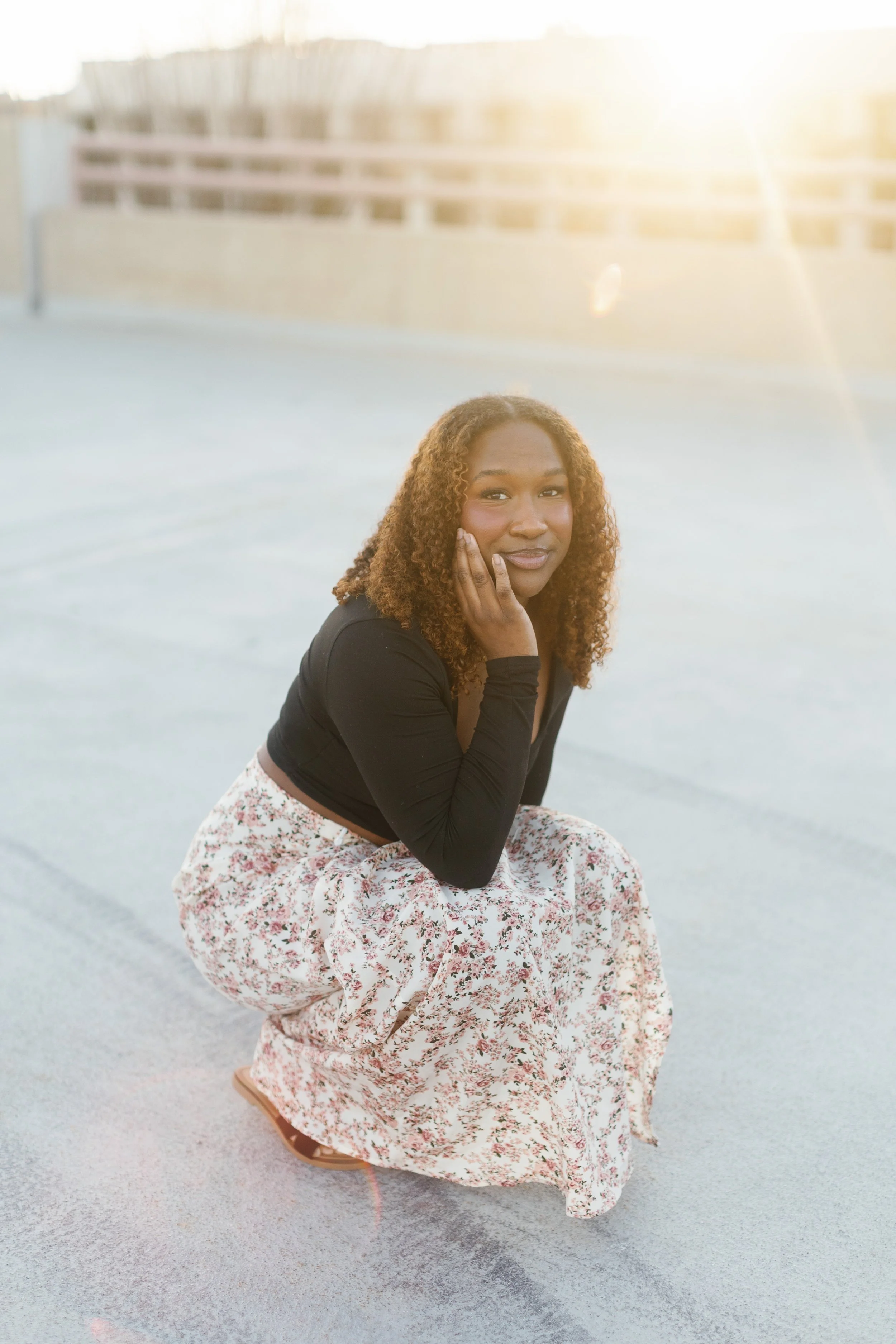 A woman kneeling on the ground with a gentle smile, one hand touching her face, wearing a black top and floral skirt, in an outdoor setting during sunset.