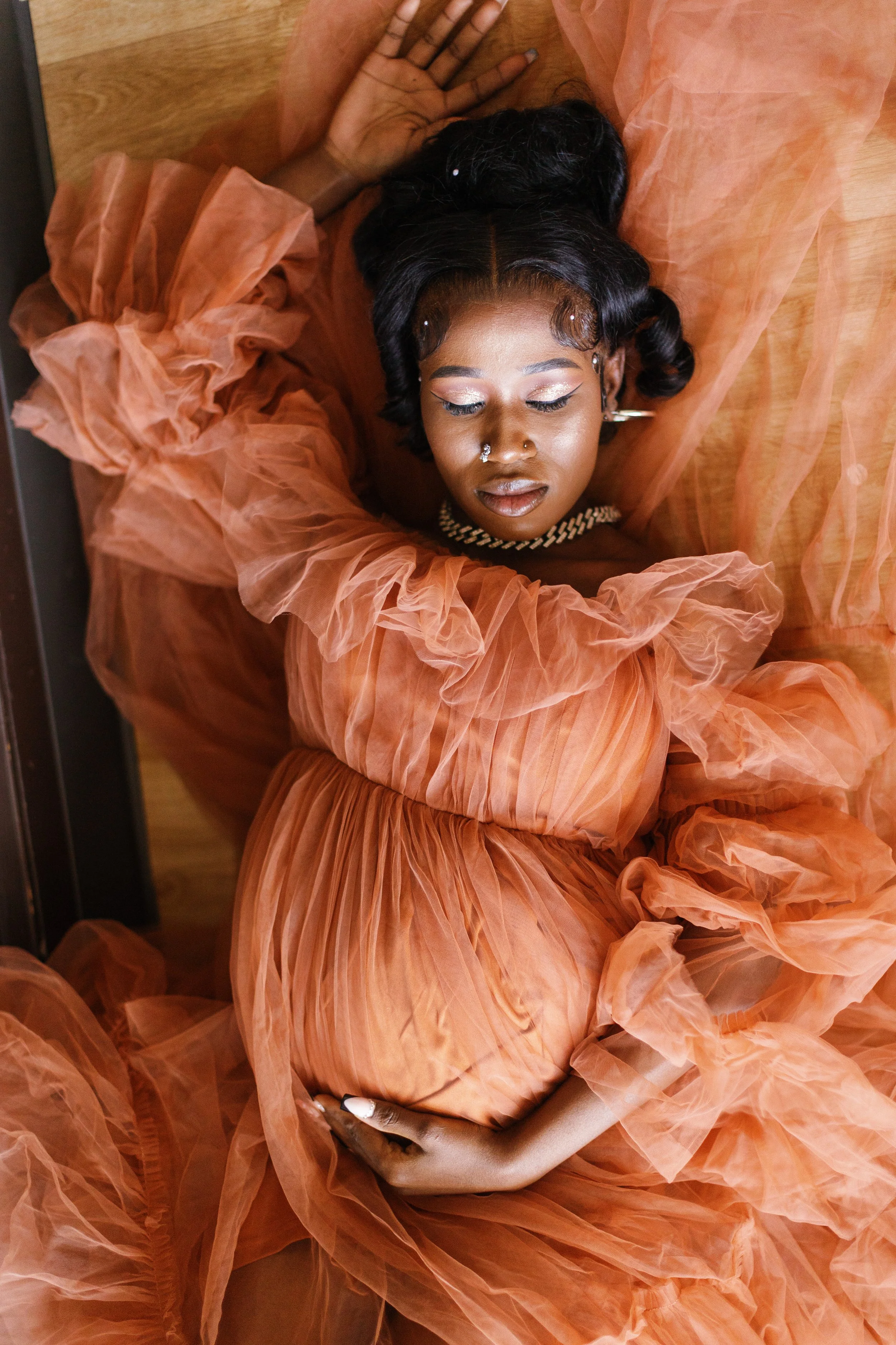 A woman lying on a wooden floor dressed in an orange, sheer, ruffled gown with makeup, jewelry, and a nose piercing, with her eyes closed.