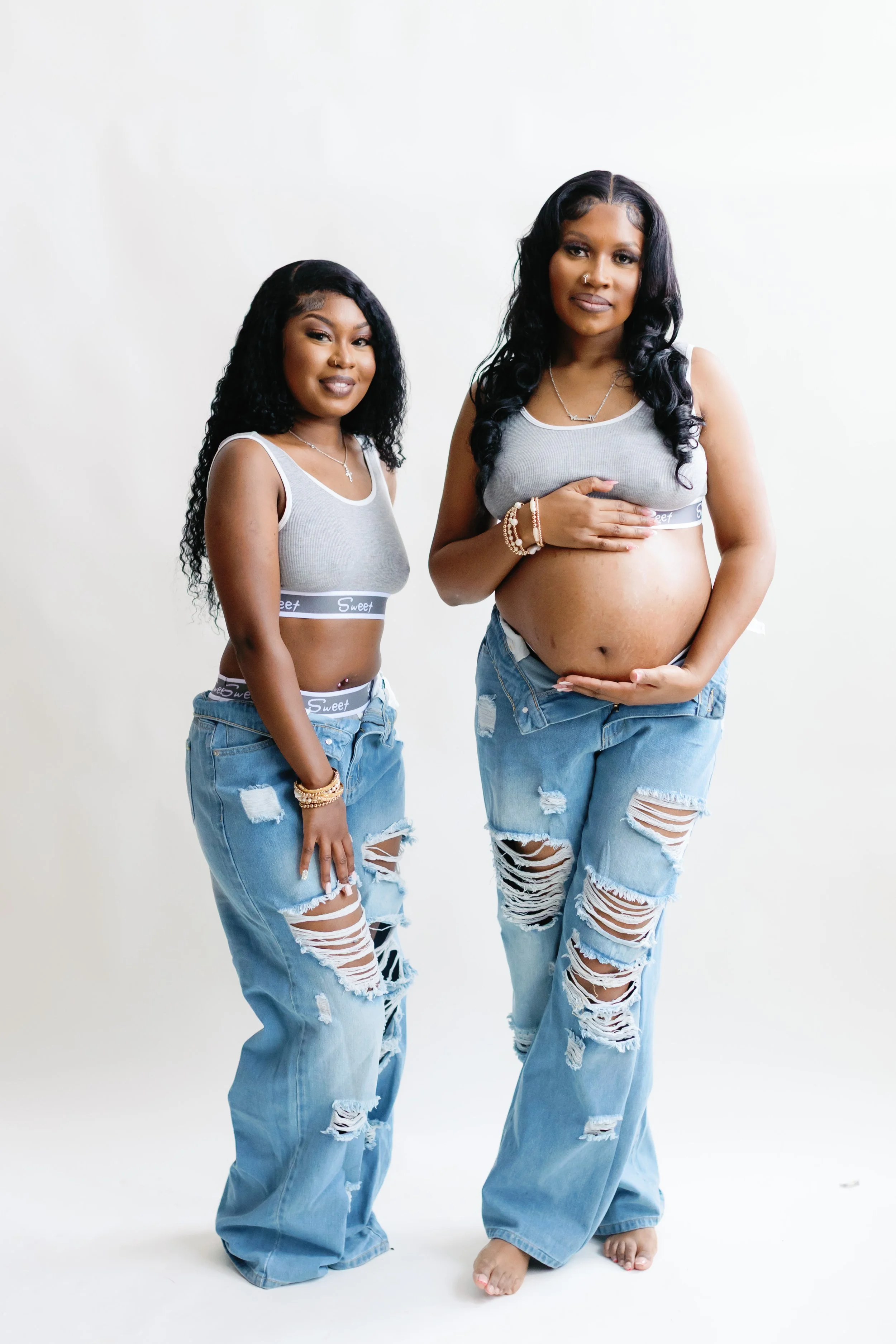Two women with long dark wavy hair posing against a white background, wearing gray crop tops, ripped jeans, and jewelry, with one pregnant woman holding her belly.
