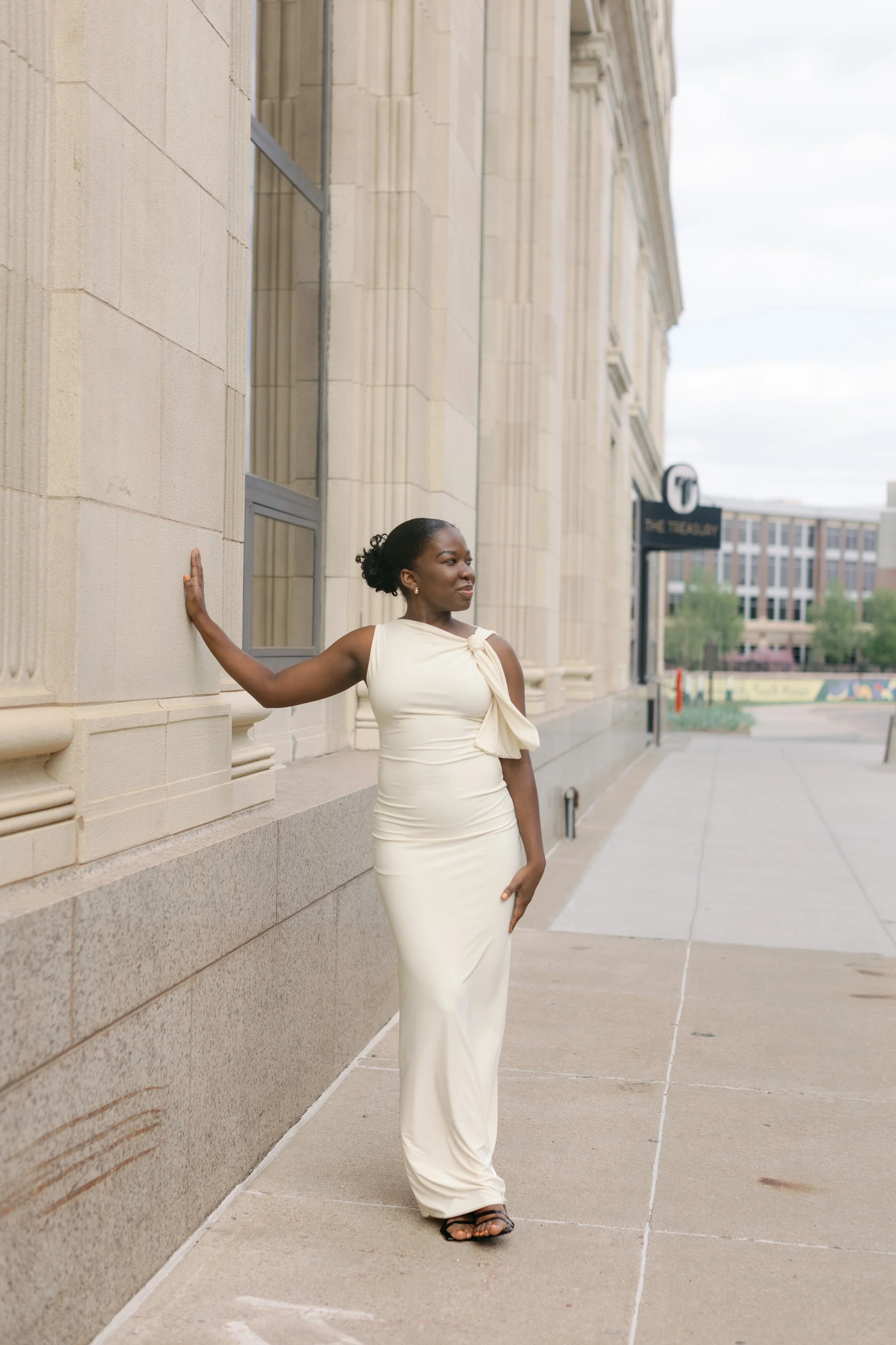 A woman in a cream-colored dress standing outdoors against a stone building wall, with her hand on the wall and looking to her right.
