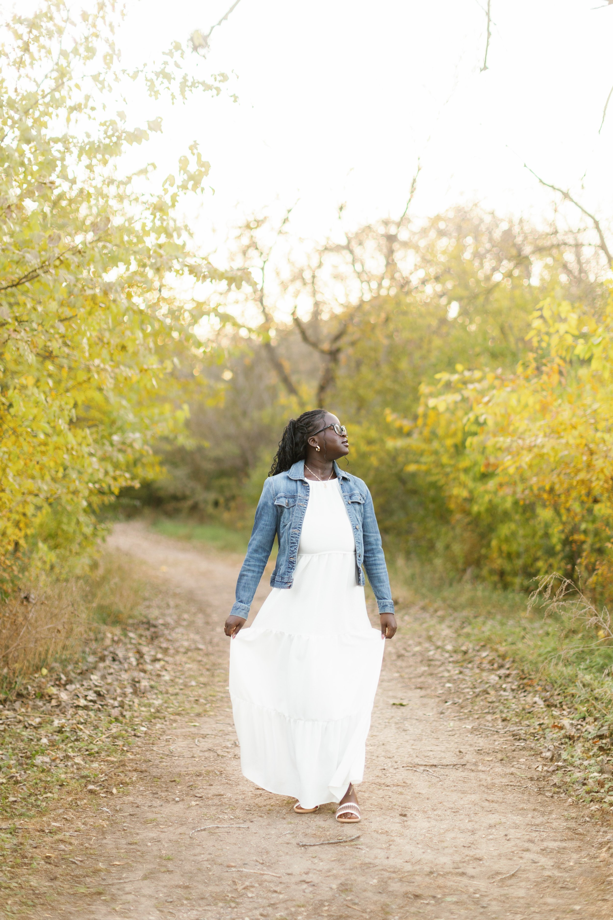 A woman wearing a white dress, a denim jacket, and glasses walking on a dirt path surrounded by trees with yellow and green leaves.