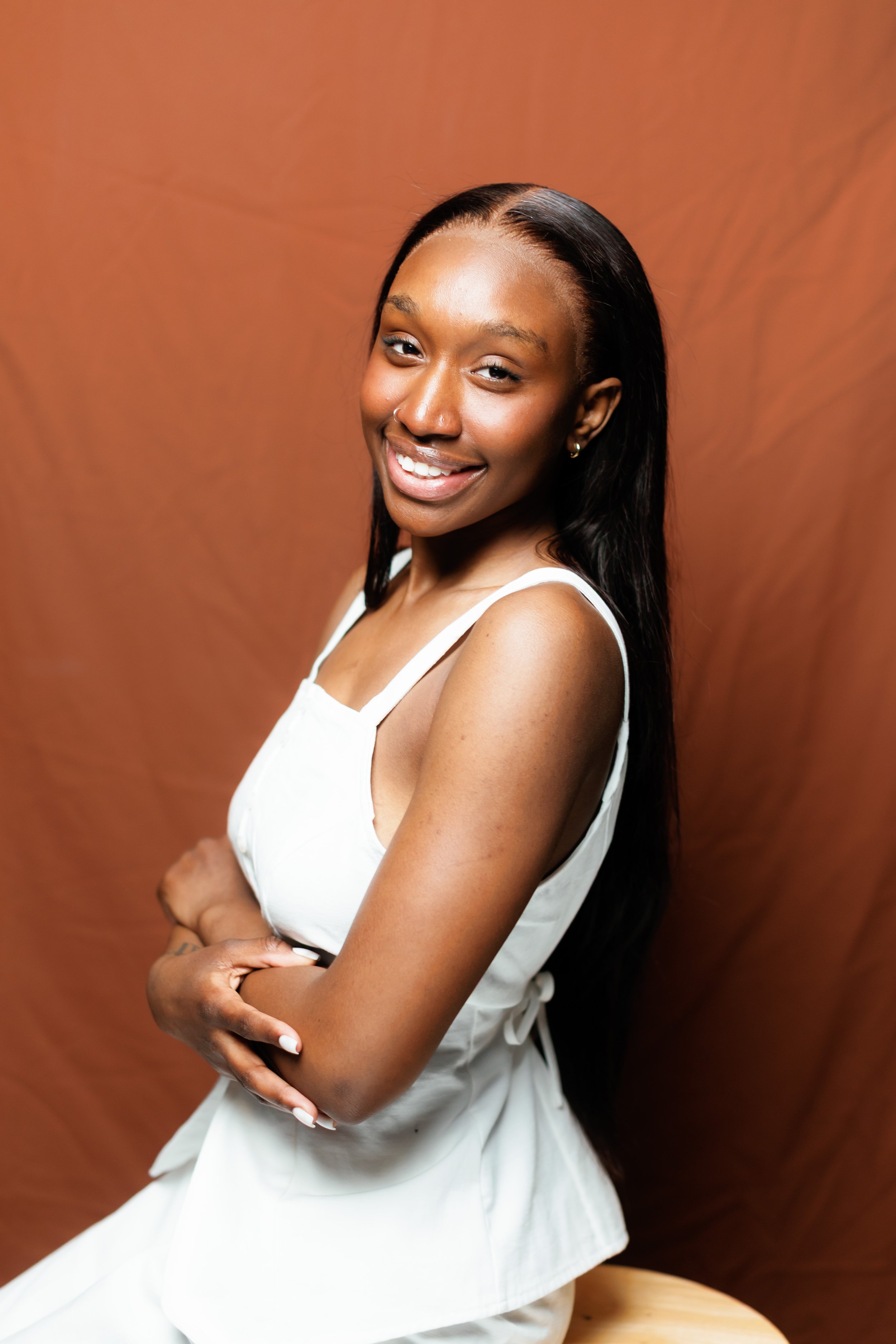 A young woman with long black hair, smiling, wearing a white sleeveless dress, standing against a plain brown background.