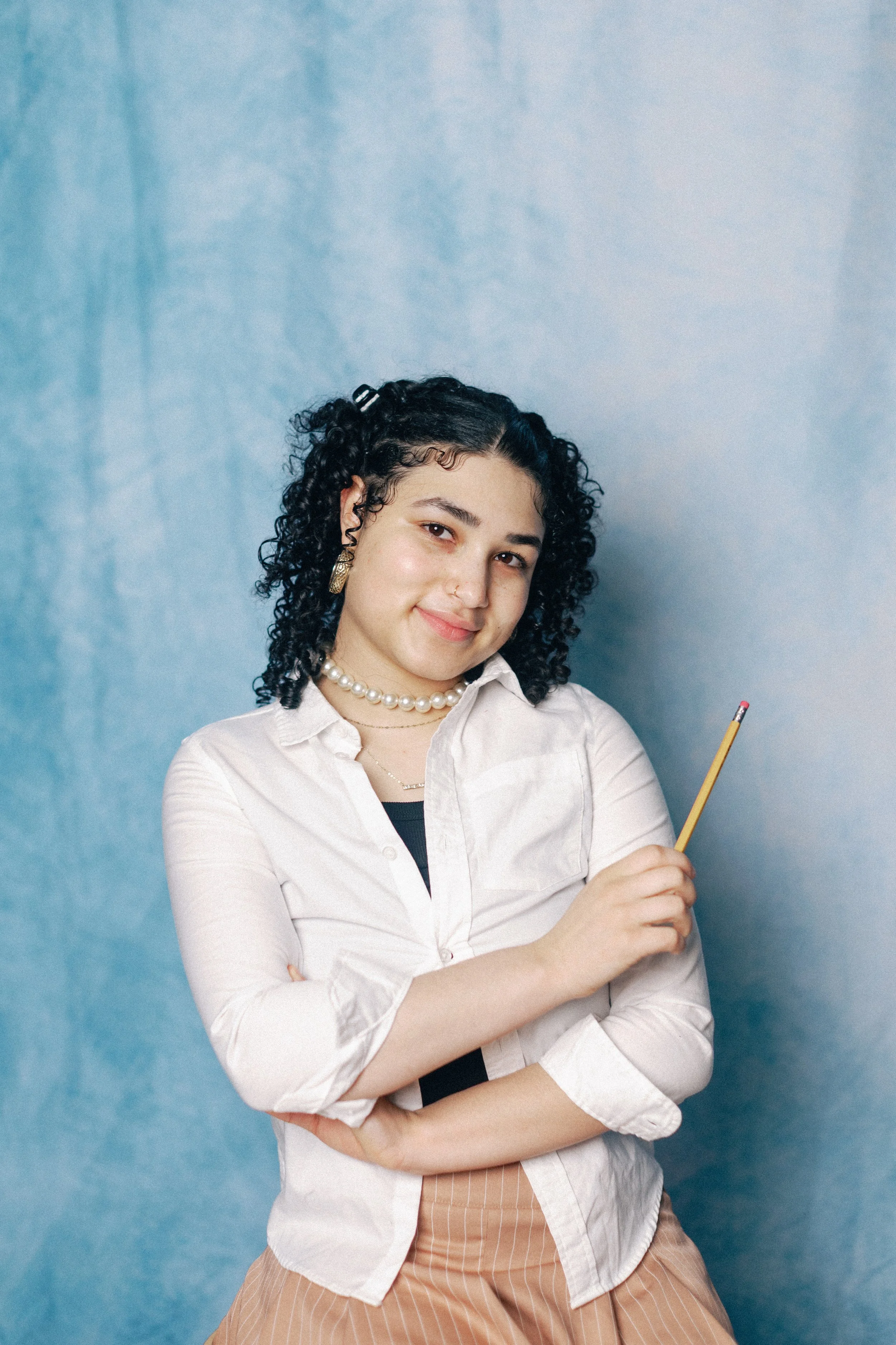 A woman with curly black hair, wearing a white shirt, pearl necklace, gold earrings, and holding a pencil, smiles against a blue textured background.