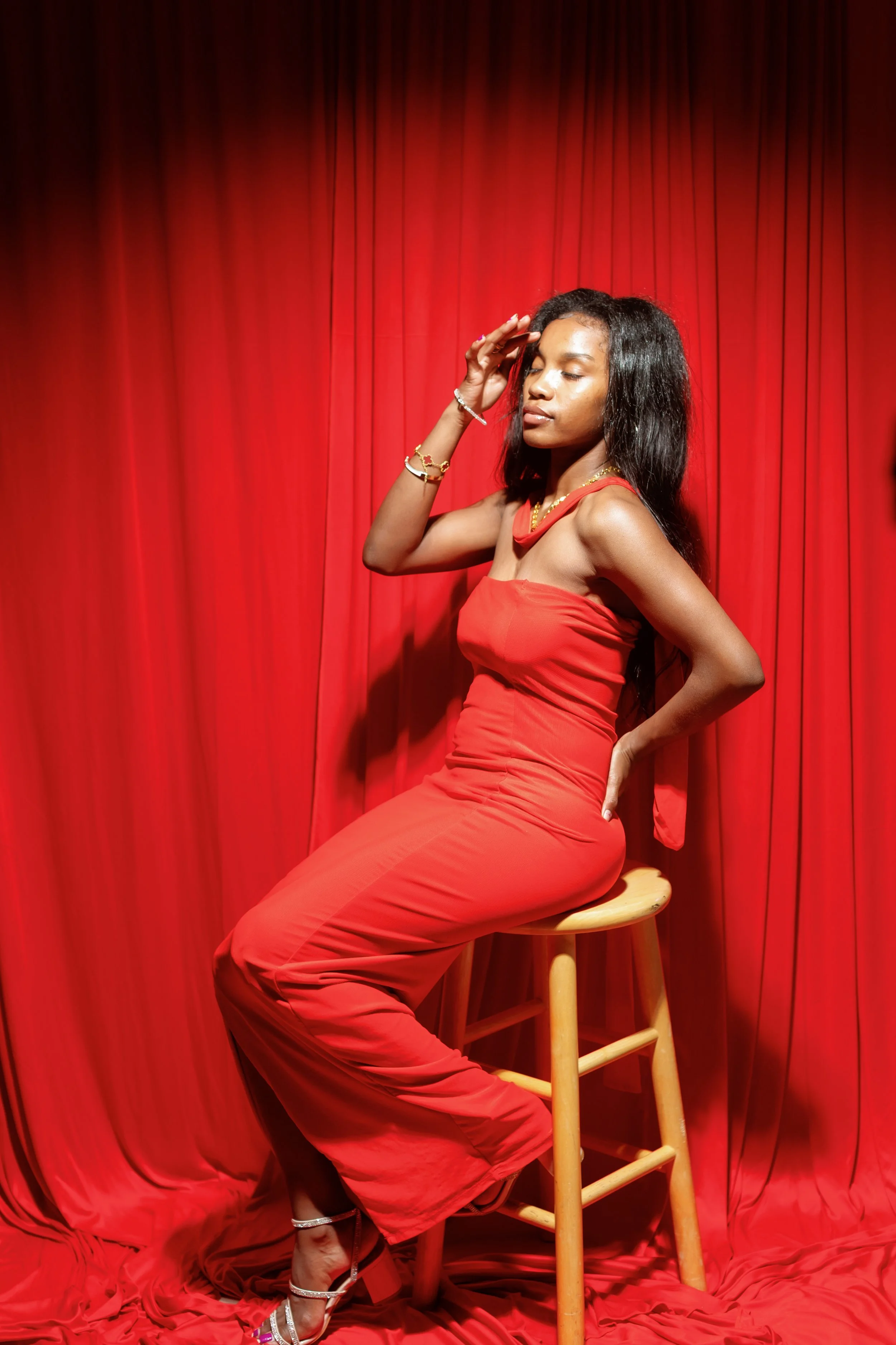 A woman in a red dress sitting on a wooden stool in front of a red curtain with her eyes closed, right hand on her forehead, and left hand on her hip.