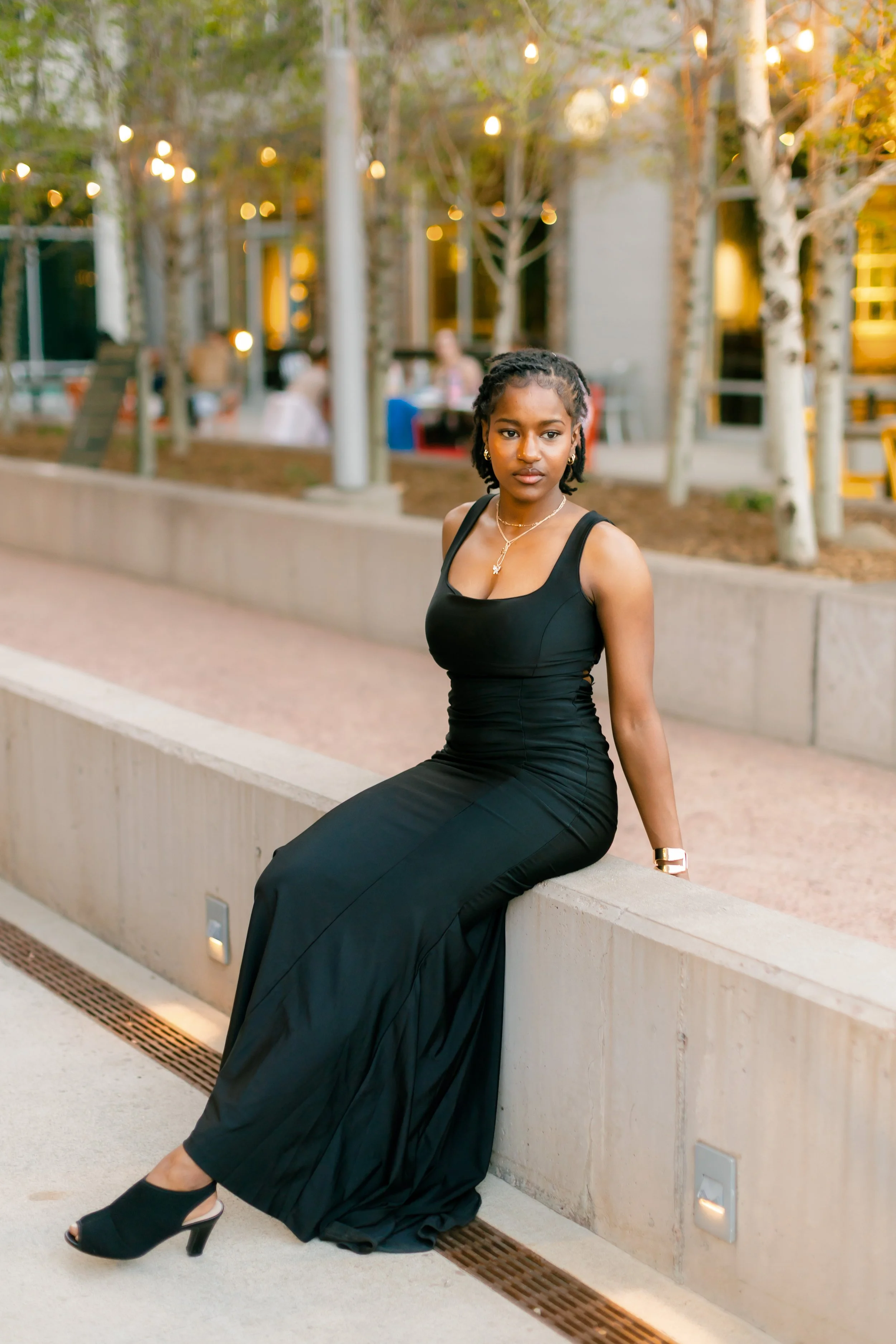A young woman with short braided hair is sitting on a concrete ledge outdoors, wearing a black sleeveless dress, black high heels, and jewelry, with blurred people and lights in the background.