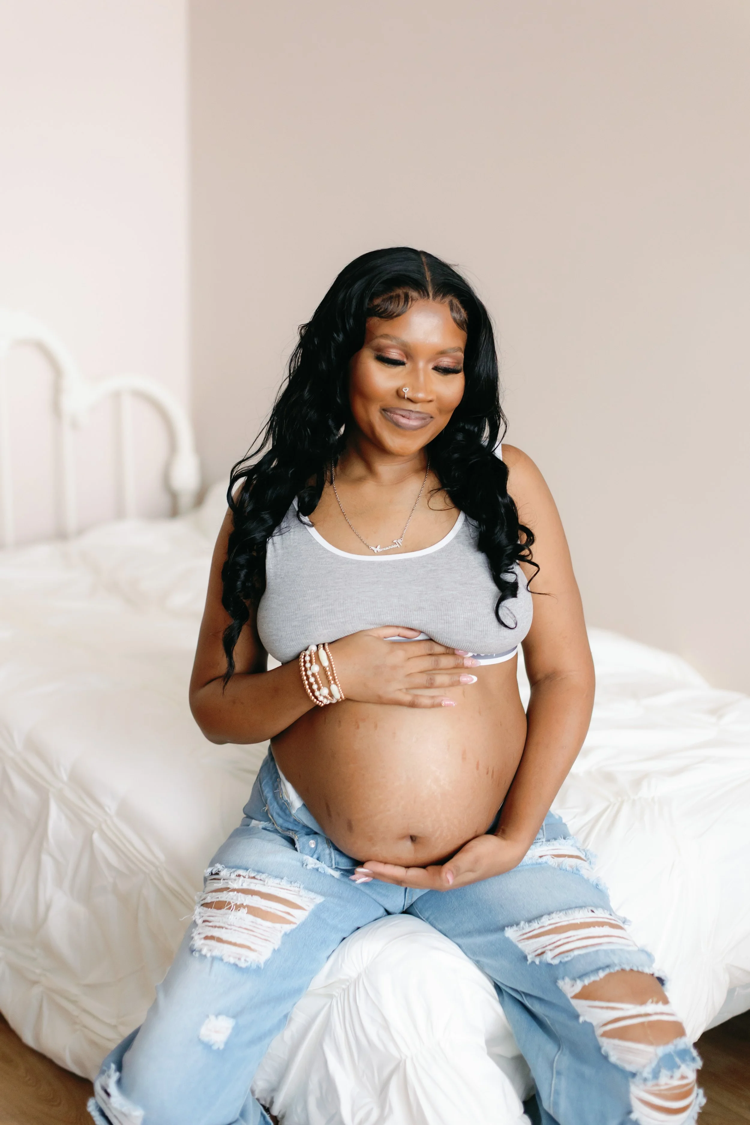 A pregnant woman with long black wavy hair and a nose piercing, smiling softly with her eyes closed, sitting on a white bed in a bedroom with beige walls. She is wearing a gray sports bra, ripped light blue jeans, and has jewelry including bracelets 