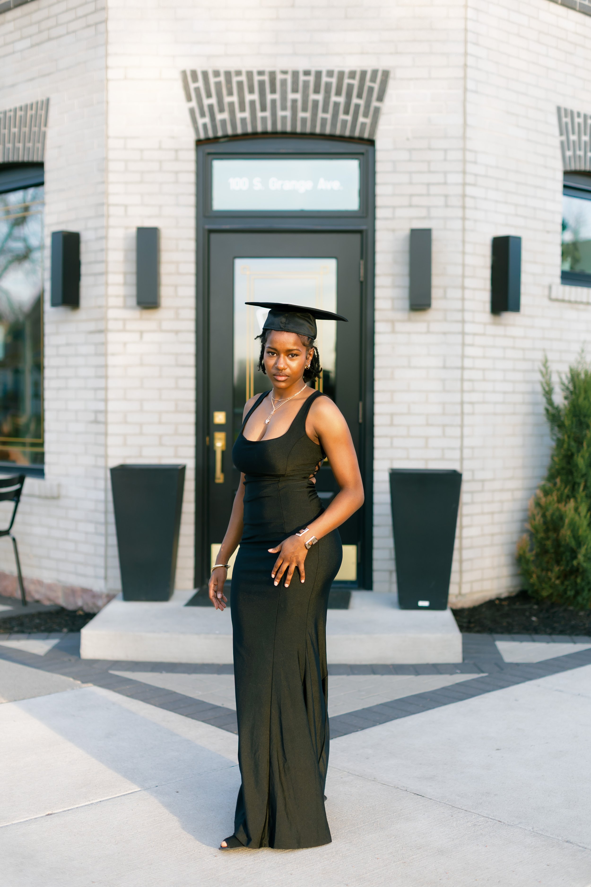Young woman in a black dress and graduation cap standing in front of a modern brick building with large windows and black planters.