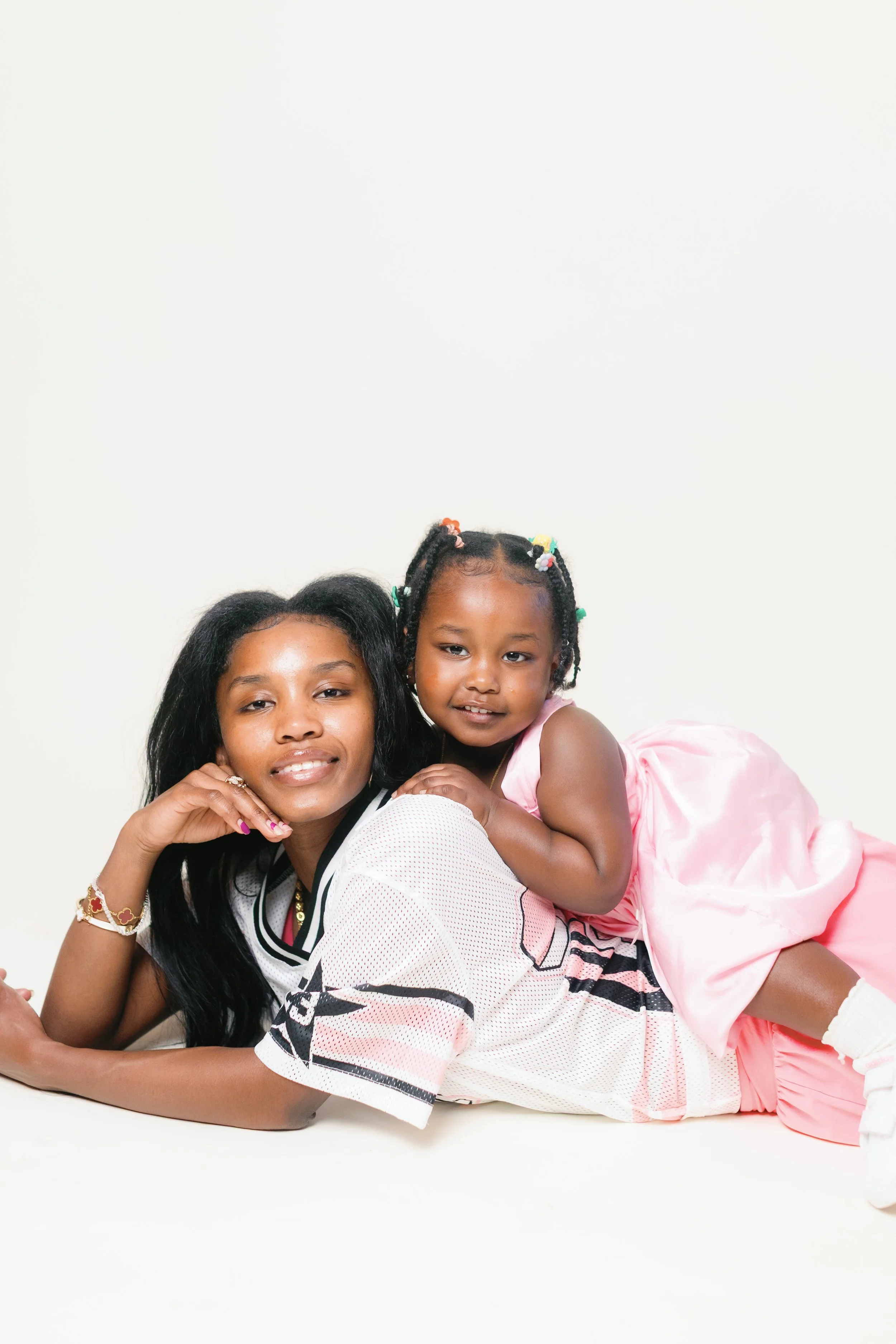 A woman and a young girl lying on the floor, smiling at the camera. The woman has long black hair and is resting her chin on her hand. The girl, wearing a pink dress with colorful hair beads, is lying on top of the woman, smiling.