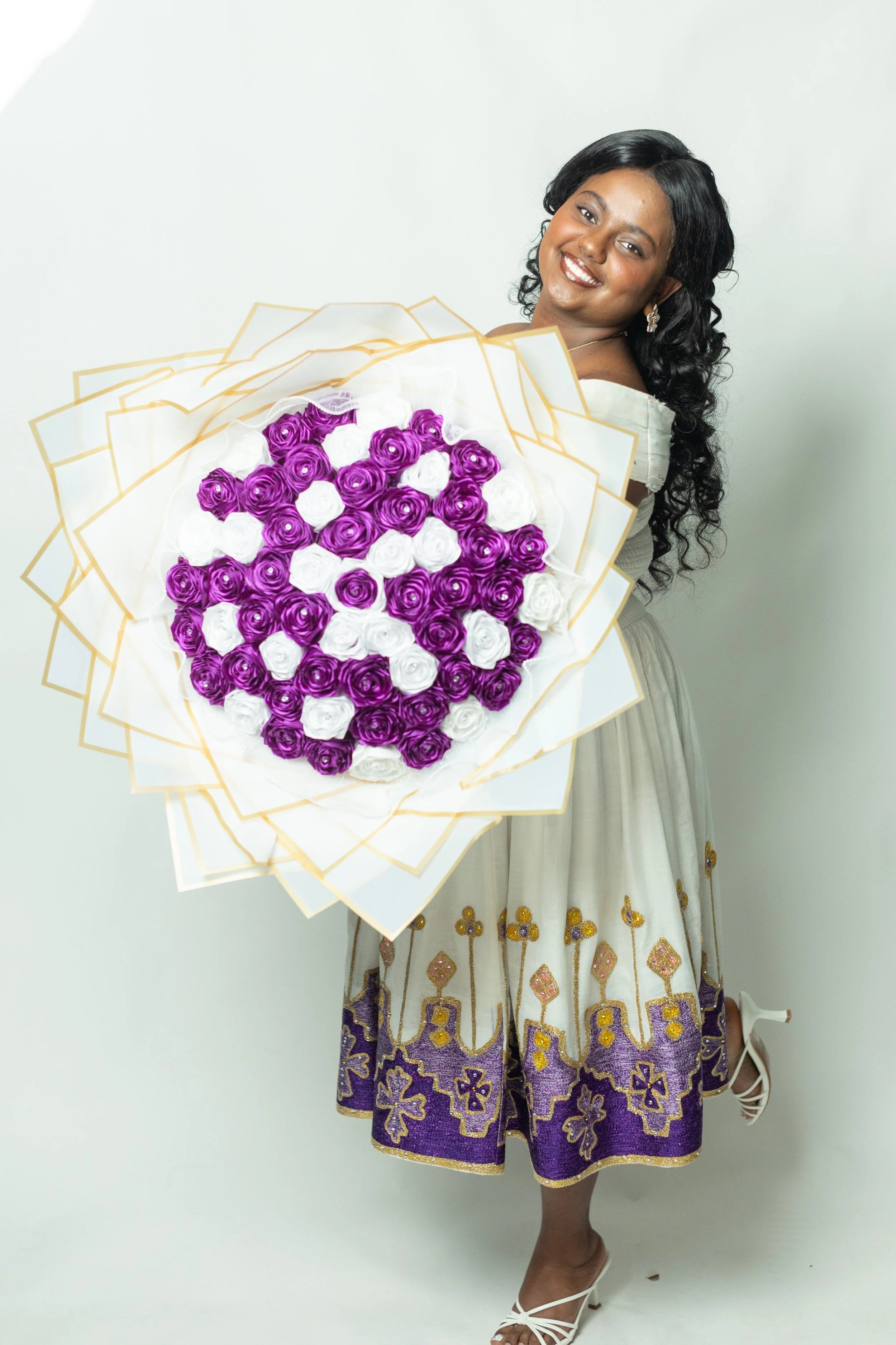 A woman with long, dark curly hair wearing a traditional dress with gold embellishments, holding a bouquet of purple and white roses, posing against a plain white background.