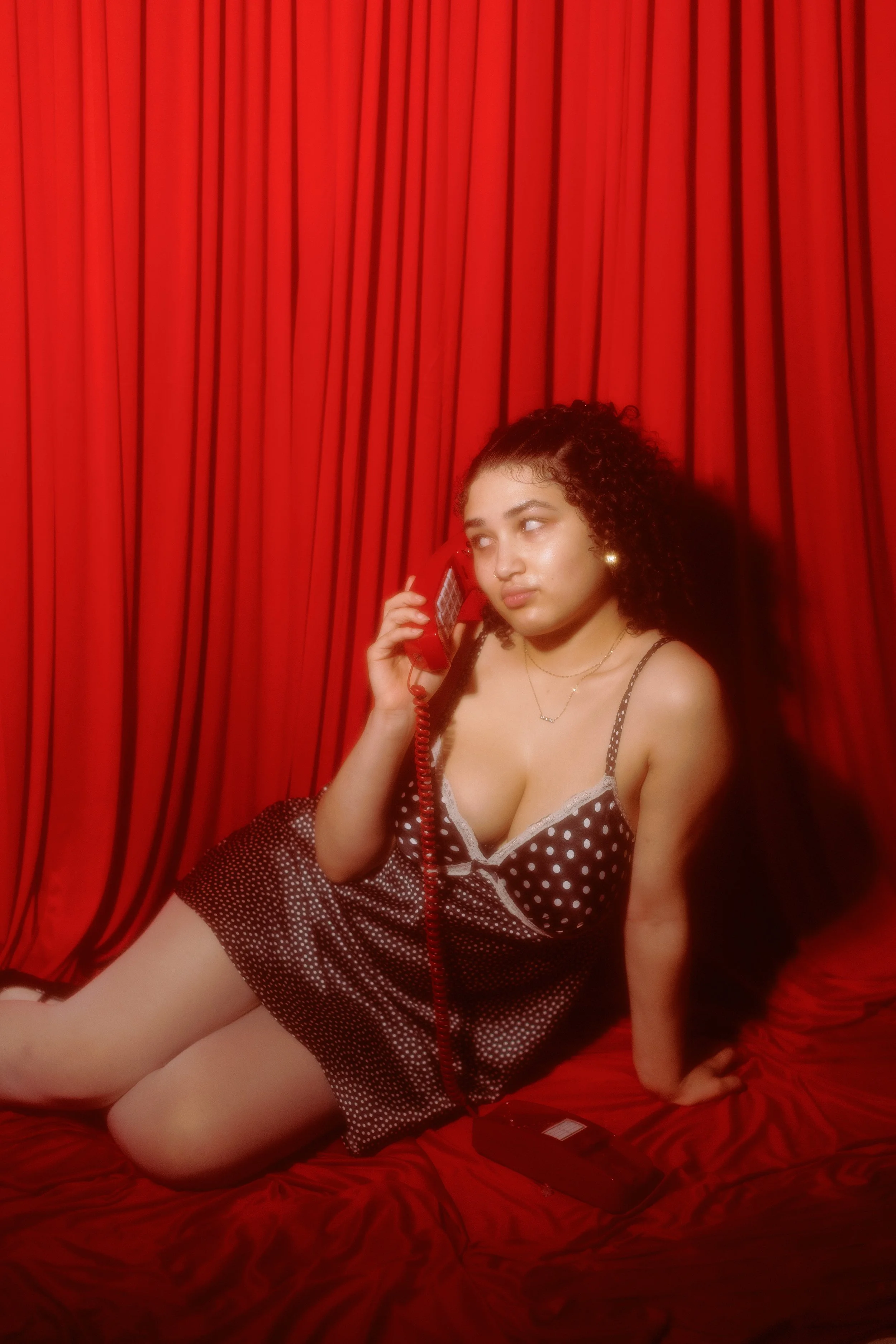 A young woman with curly hair wearing polka dot pajamas, sitting on a red bed with red curtains behind her, talking on a red rotary phone.