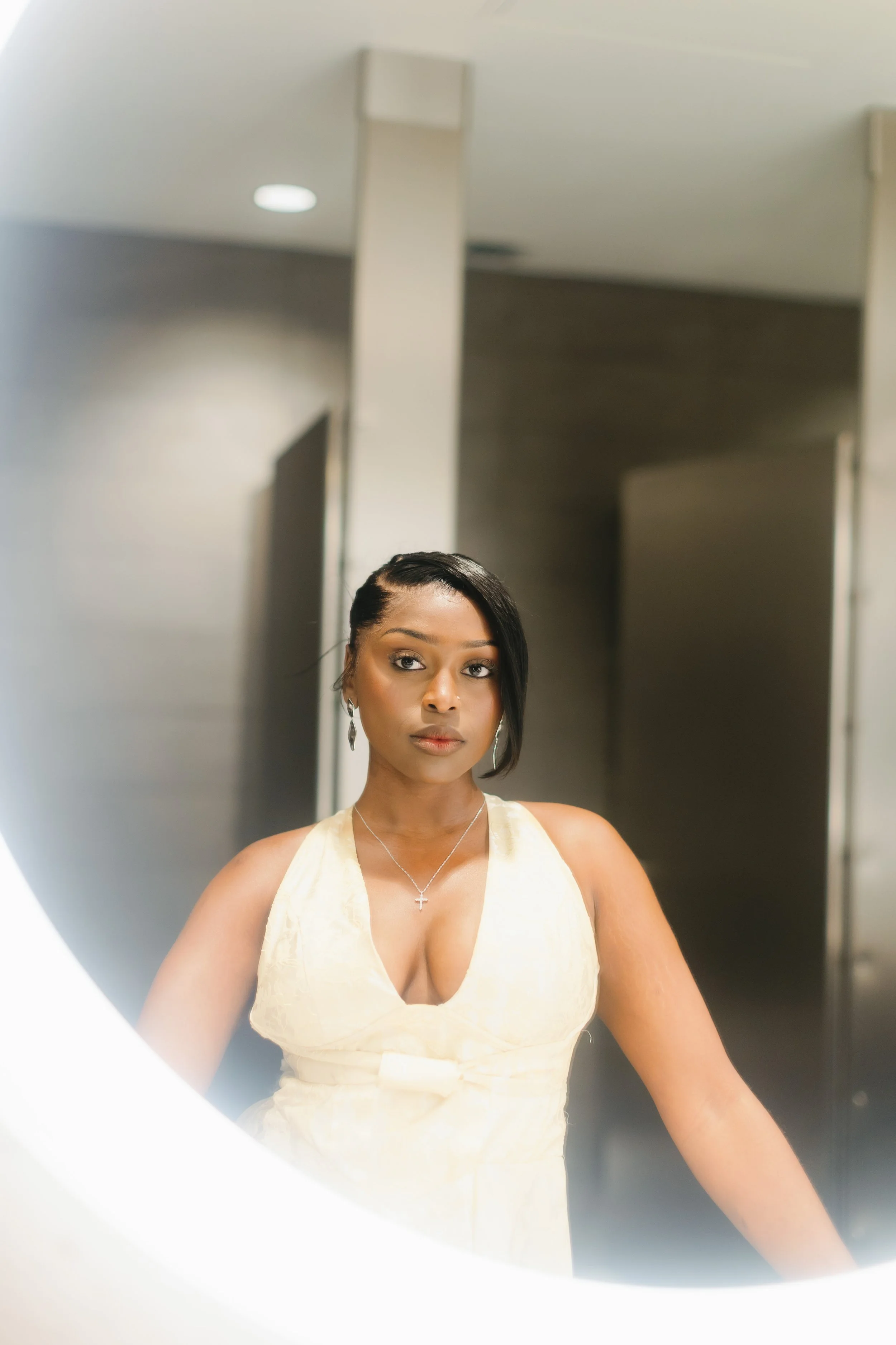 A woman with dark hair styled in a side part, wearing a light yellow dress with a deep V-neckline, earrings, and a cross necklace, taking a selfie in a mirror.