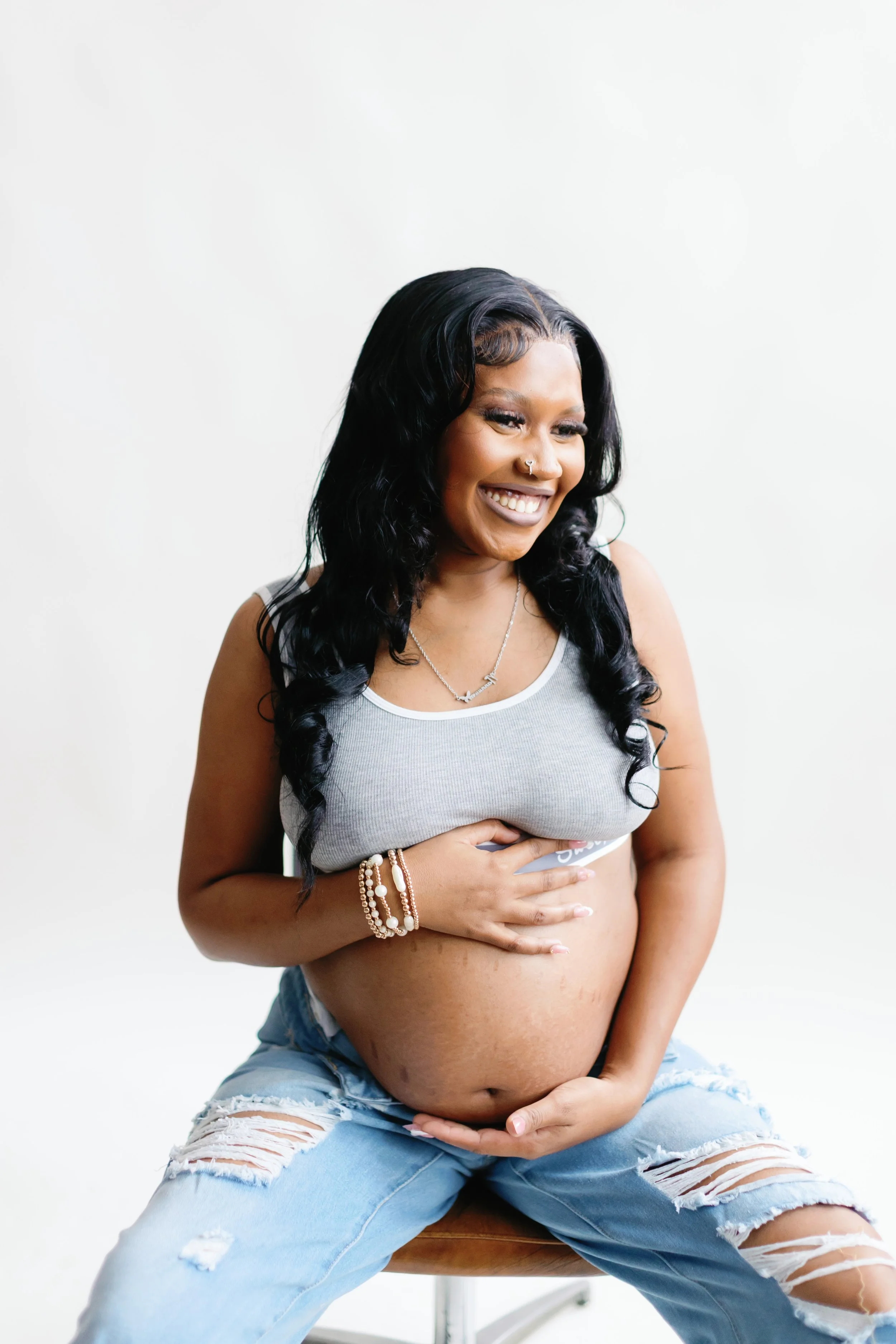 A pregnant woman with long black hair, smiling, wearing a gray crop top and ripped jeans, holding her belly with both hands, seated on a stool against a plain white background.
