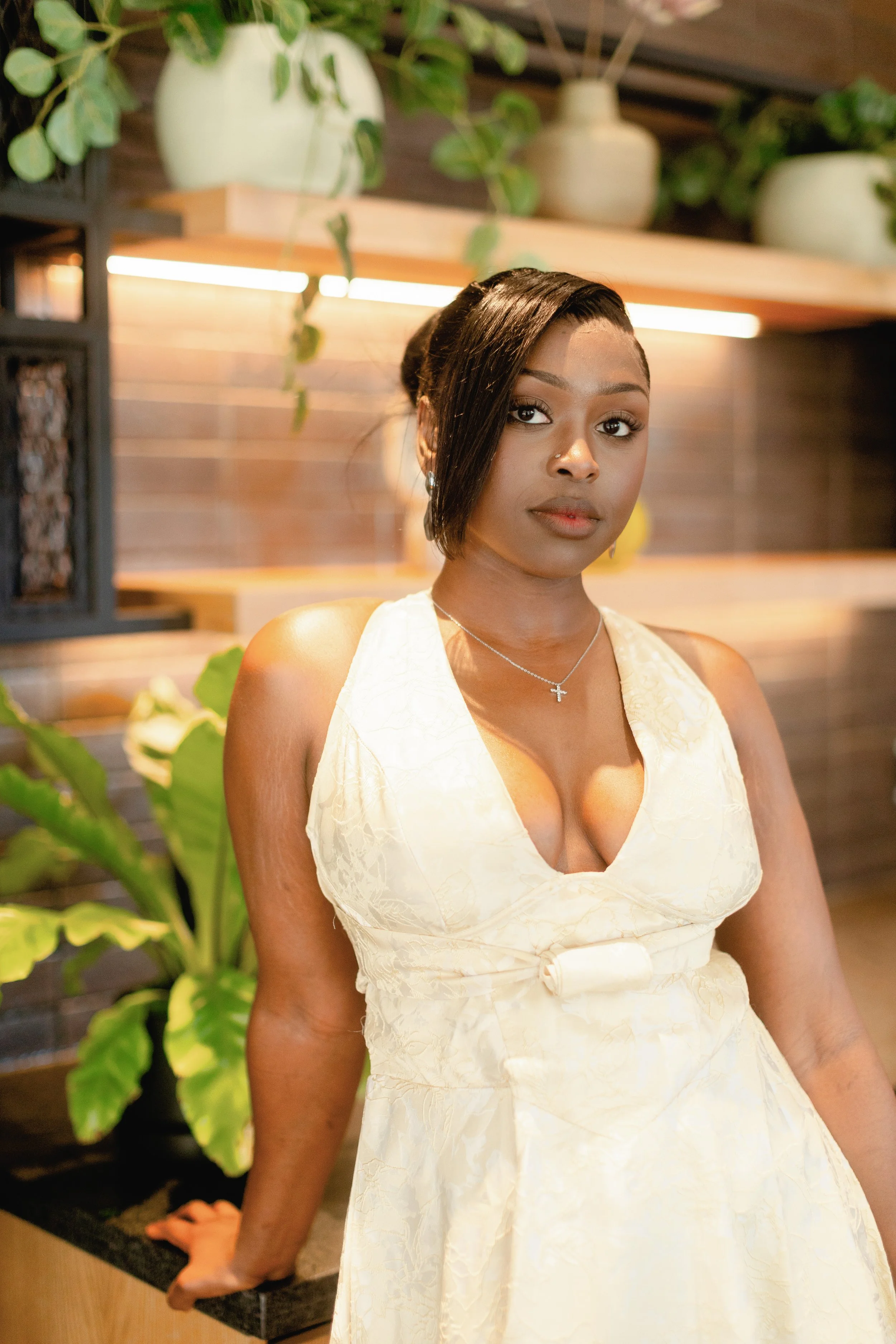 A woman with short black hair, wearing a cream-colored dress and a silver cross necklace, standing indoors in front of a wooden and brick wall with greenery and potted plants.