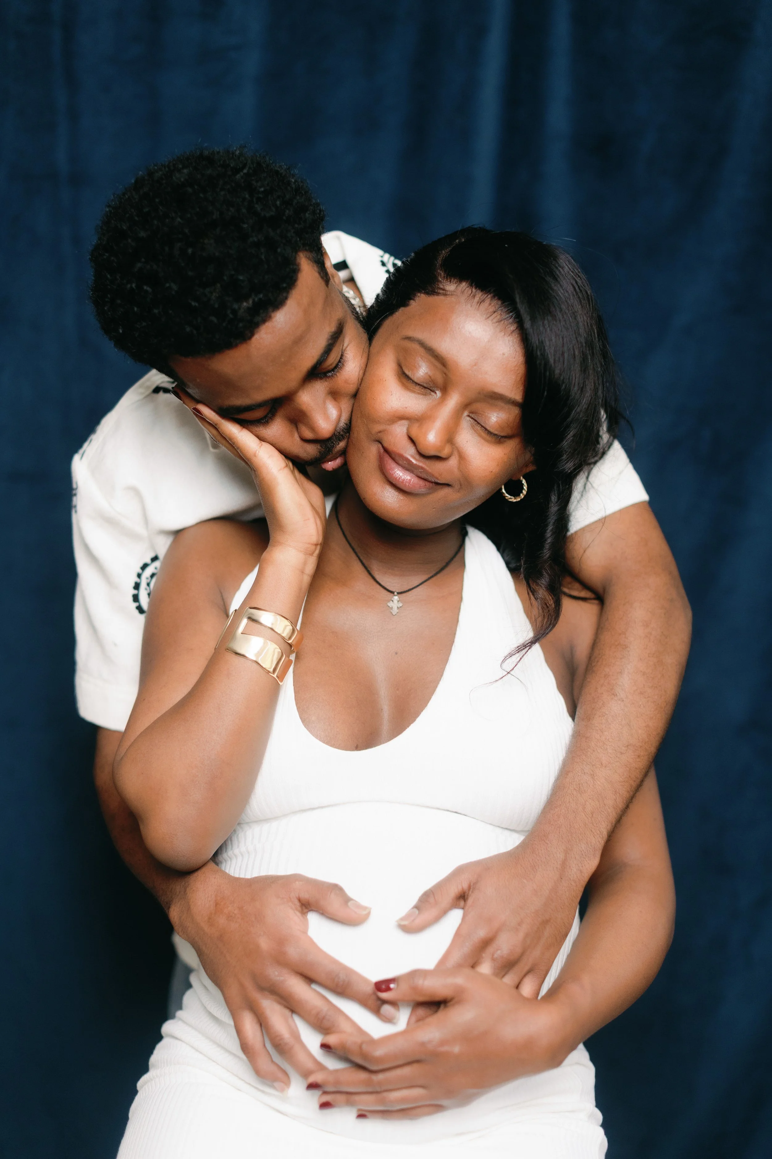A couple embracing, with the man kissing the woman's cheek and both forming a heart shape with their hands over her pregnant belly, against a dark blue background.