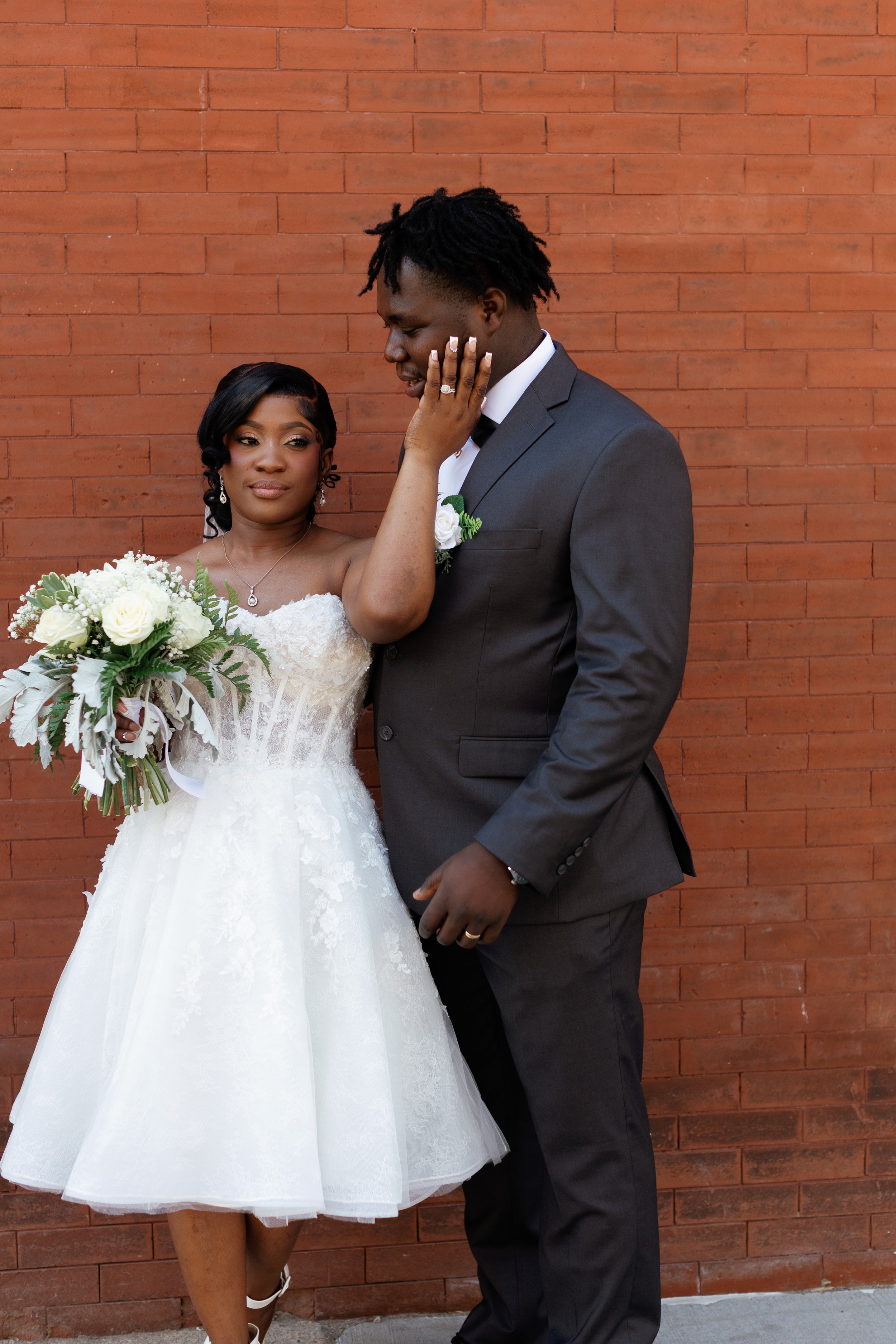 A bride and groom pose against a red brick wall. The bride is wearing a white wedding dress, holding a bouquet of white flowers, and gently touching the groom's face. The groom is dressed in a dark suit and white shirt, looking at the bride.