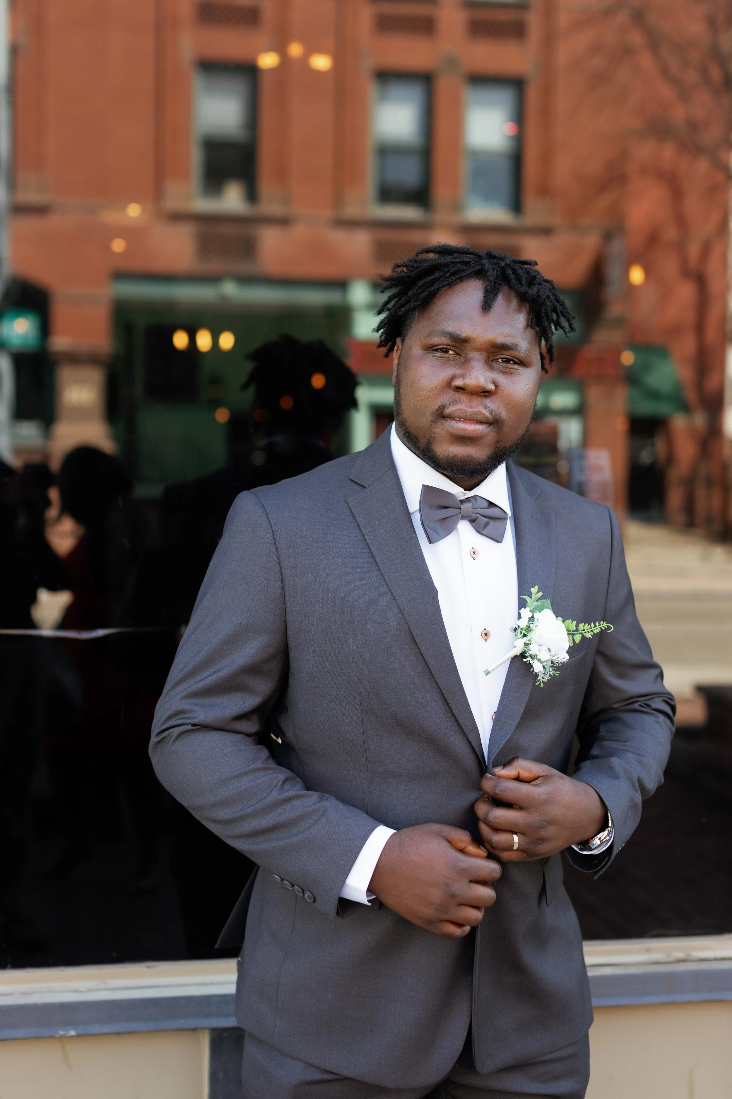 A man in a gray suit with a bow tie and boutonniere poses outdoors with a building in the background.