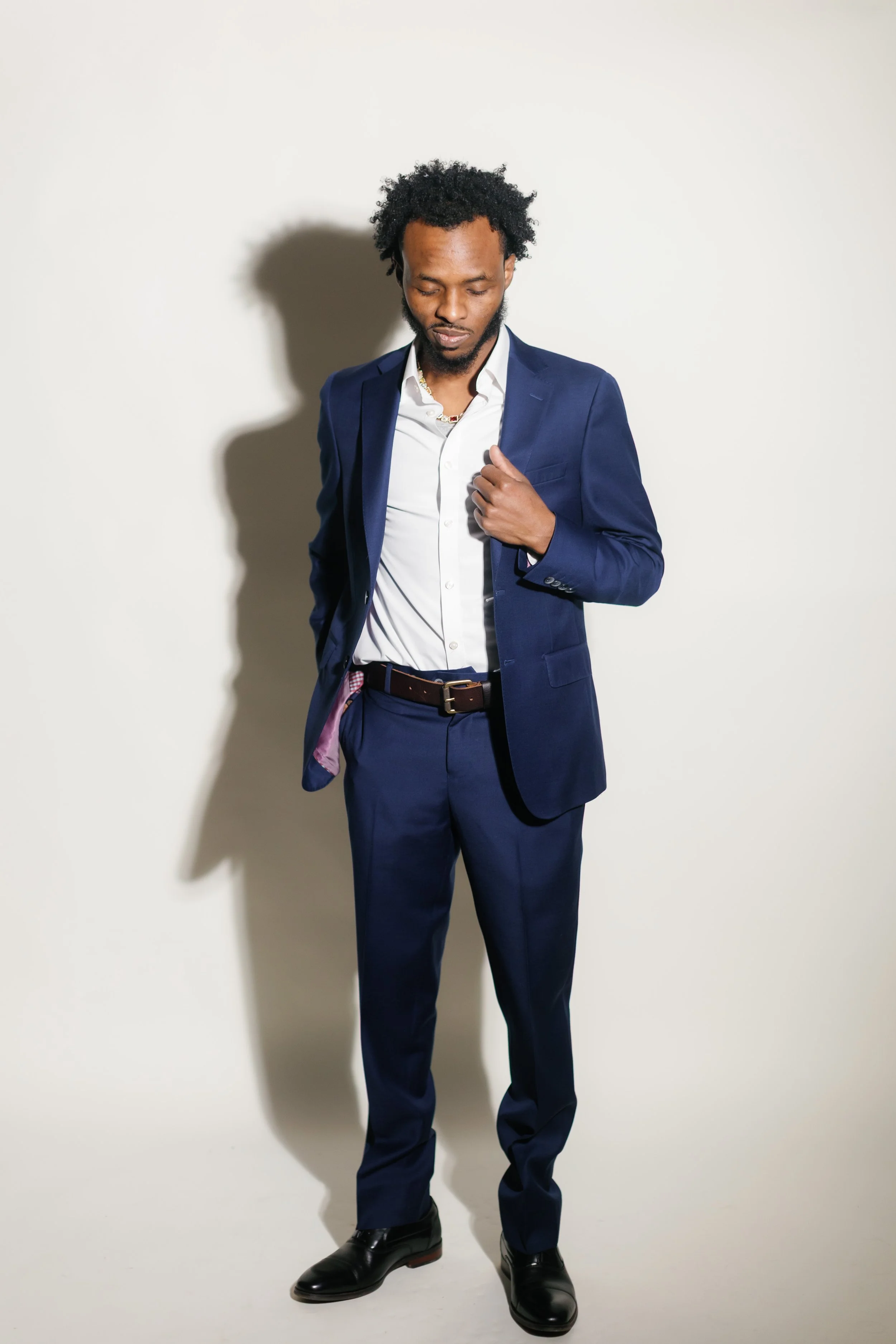 A man with curly black hair and a beard wearing a dark blue suit, white shirt, and black shoes, standing against a plain off-white background, with his eyes closed and hand adjusting his suit jacket.