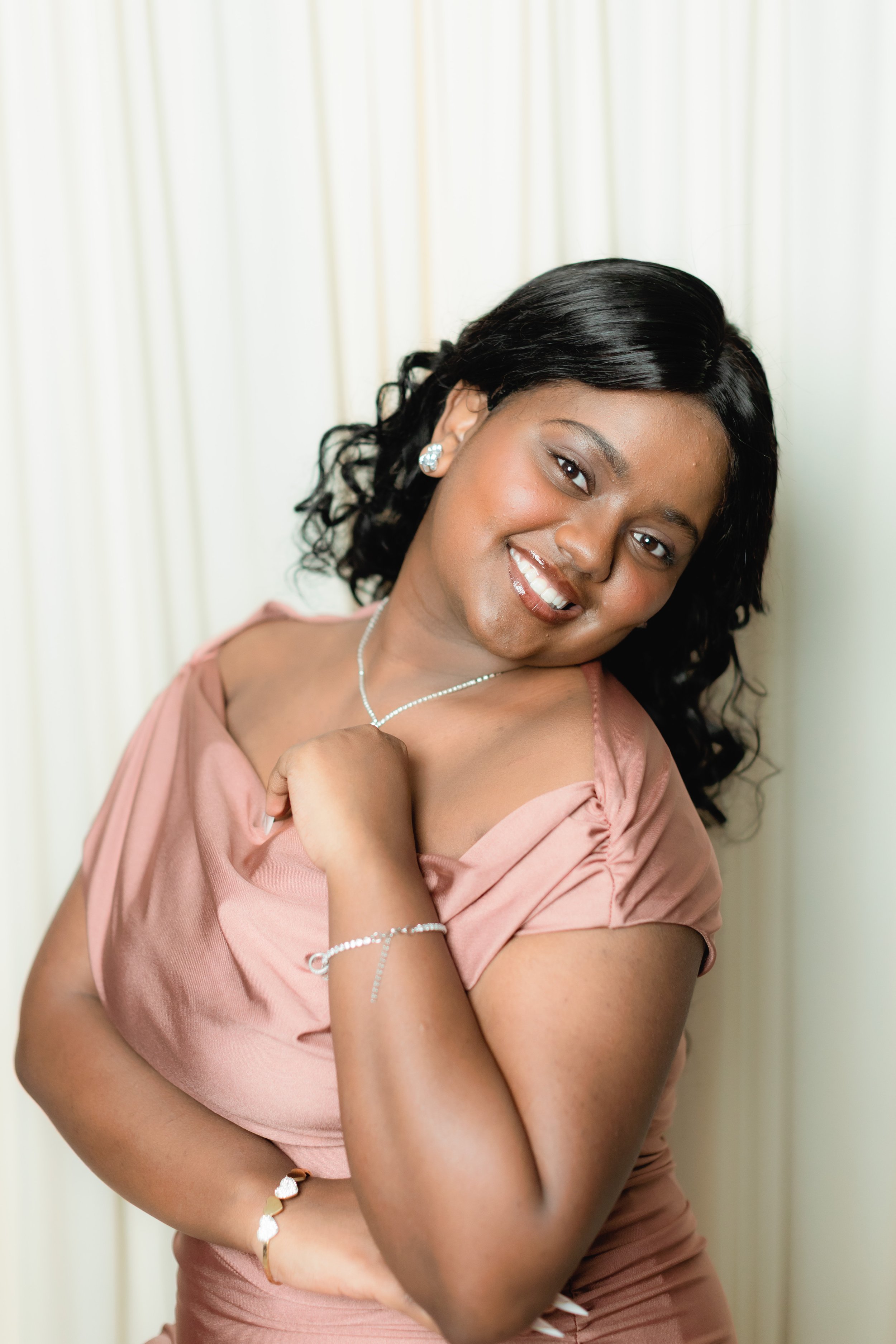 Portrait of a young woman with dark, curly hair, smiling, wearing a pink dress and jewelry, posing with her arm crossed against a neutral background.