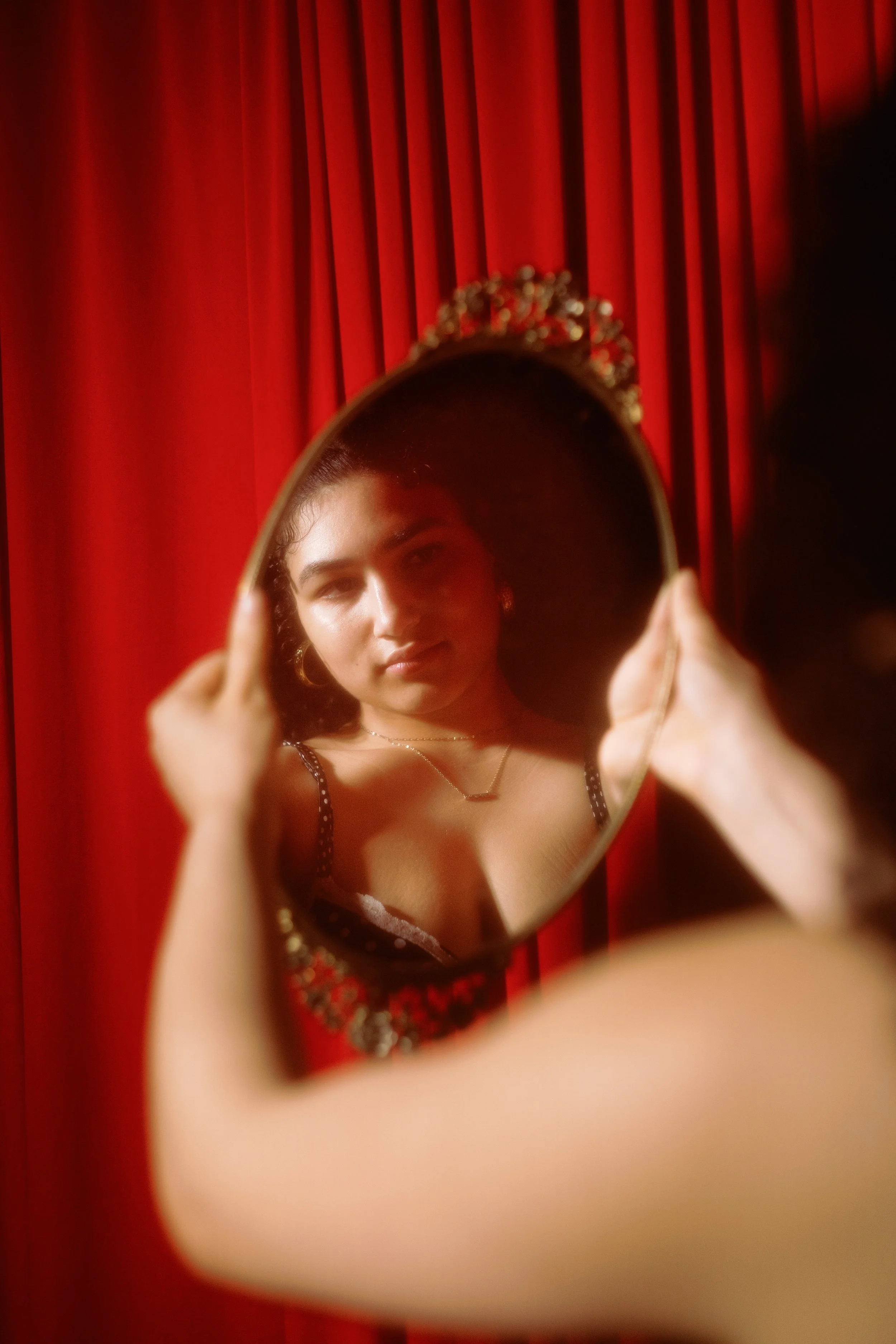 A woman looking at herself in a mirror she is holding, with red curtains in the background. She is wearing a black and red dress with spaghetti straps, and a necklace with a small bar pendant. A tiara is placed on her head. The mirror captures her fa