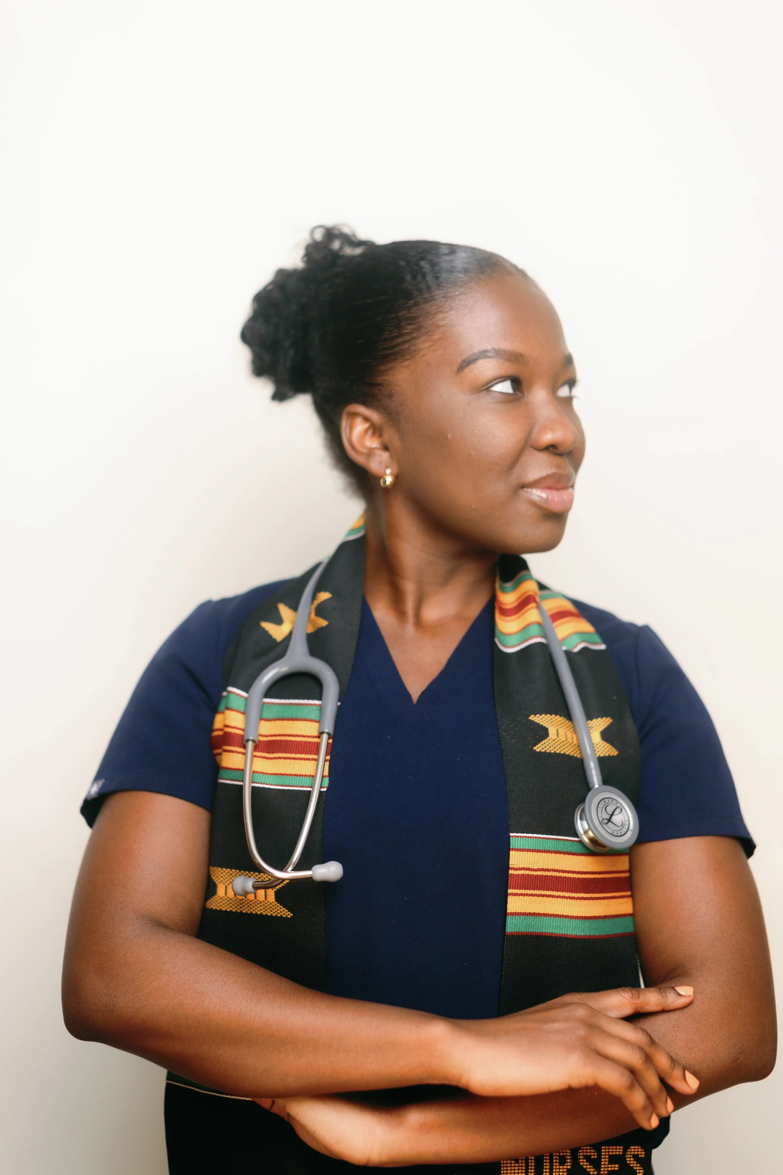 A woman with dark skin, short black hair, and gold earrings wearing a dark blue top and a colorful African-patterned scarf, with a stethoscope around her neck, standing against a plain white background.