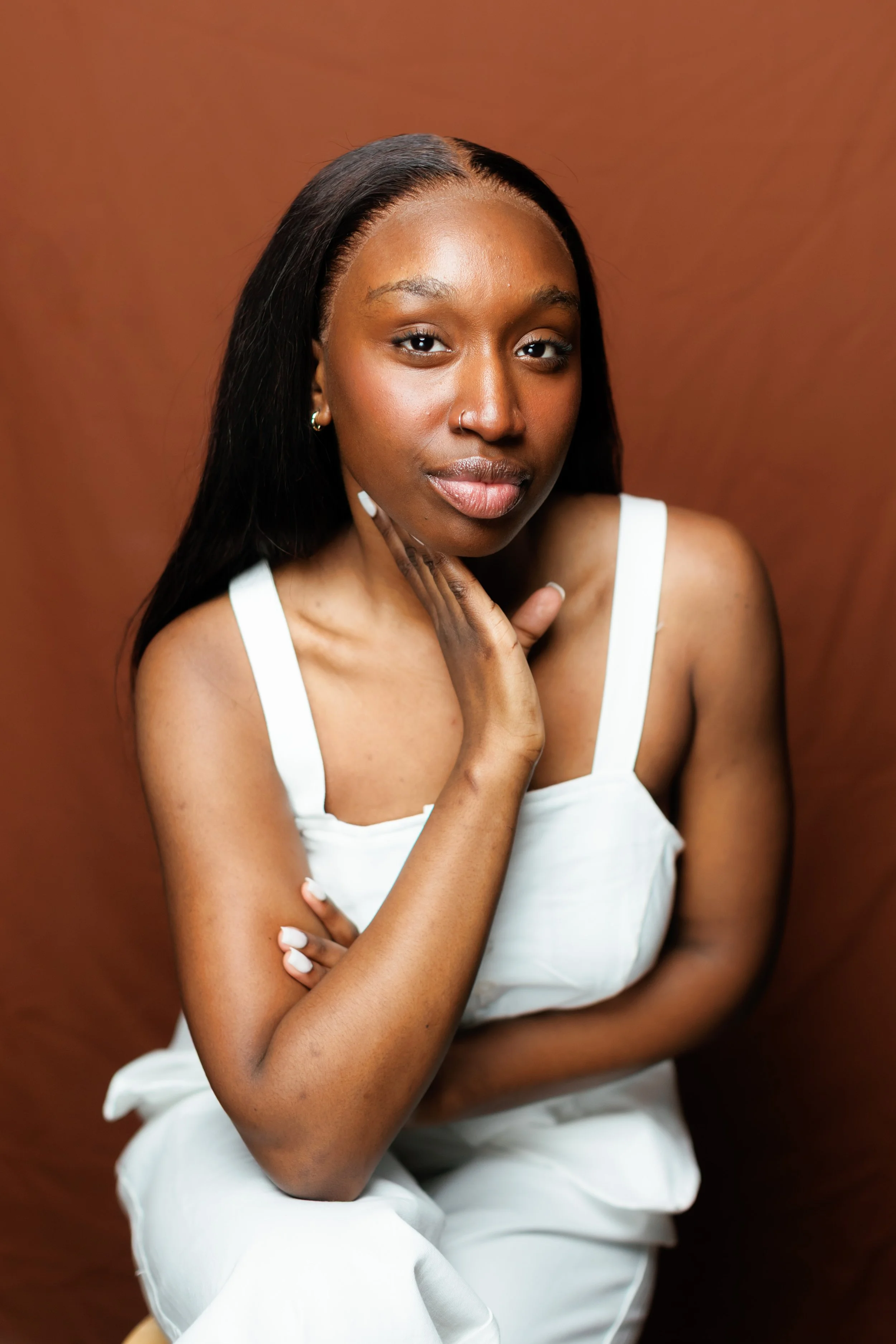 A portrait of a young woman with long black hair and dark skin, wearing a white top, with her hand near her chin, against a brown background.