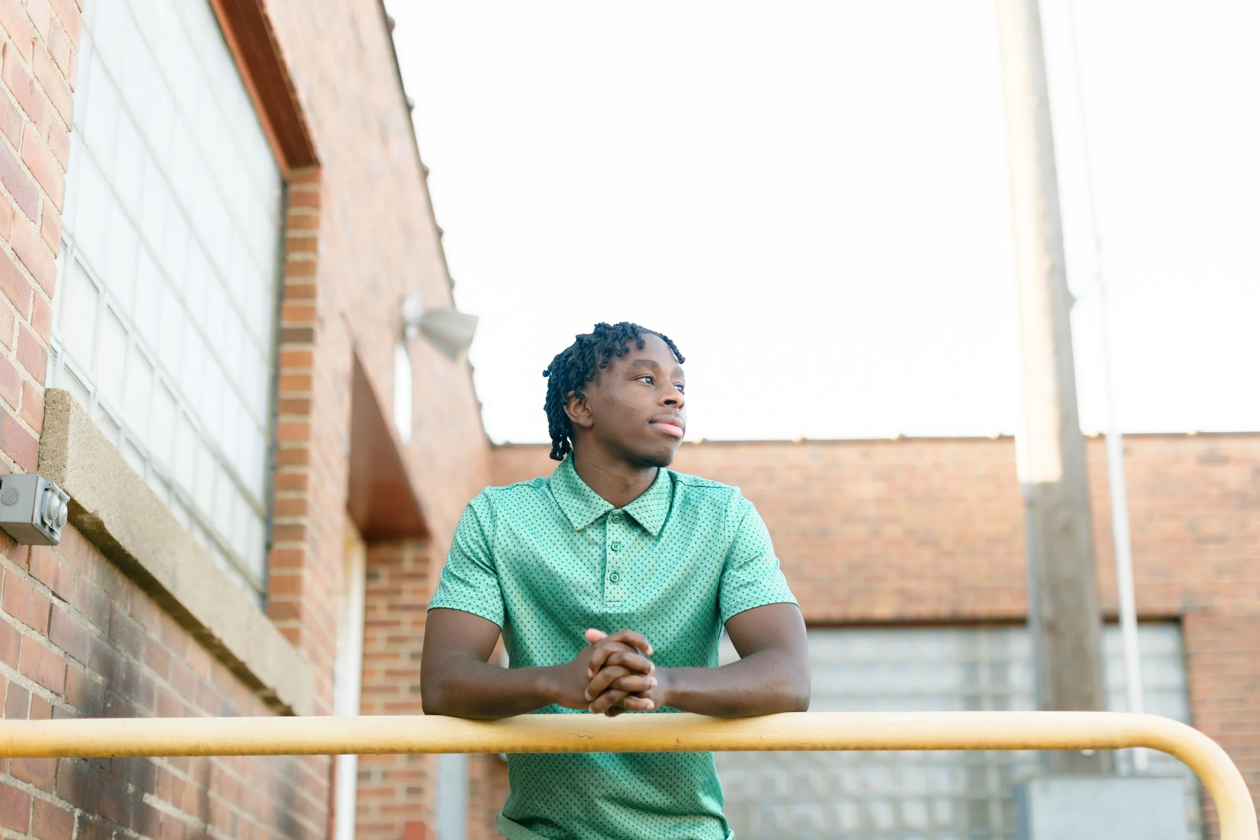 A young man with dreadlocks sitting outdoors at a table, wearing a green polka dot shirt, with arms crossed and looking to the side.