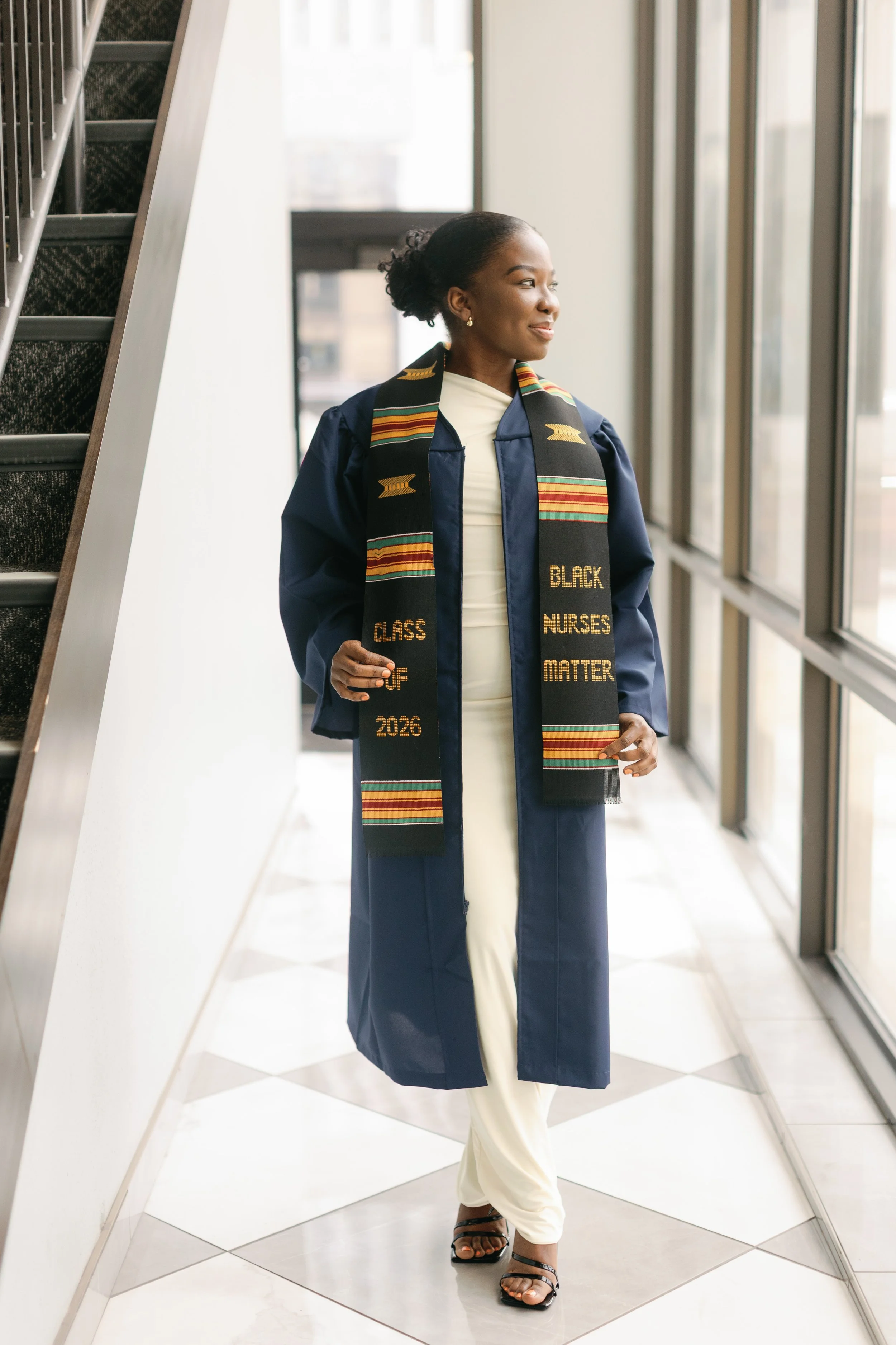 A woman in graduation attire stands indoors near large windows, wearing a dark blue gown, a colorful stole with 'Class of 2026' and 'Black Nurses Matter' embroidered on it, white pants, black sandals, and stud earrings, looking thoughtfully out the w