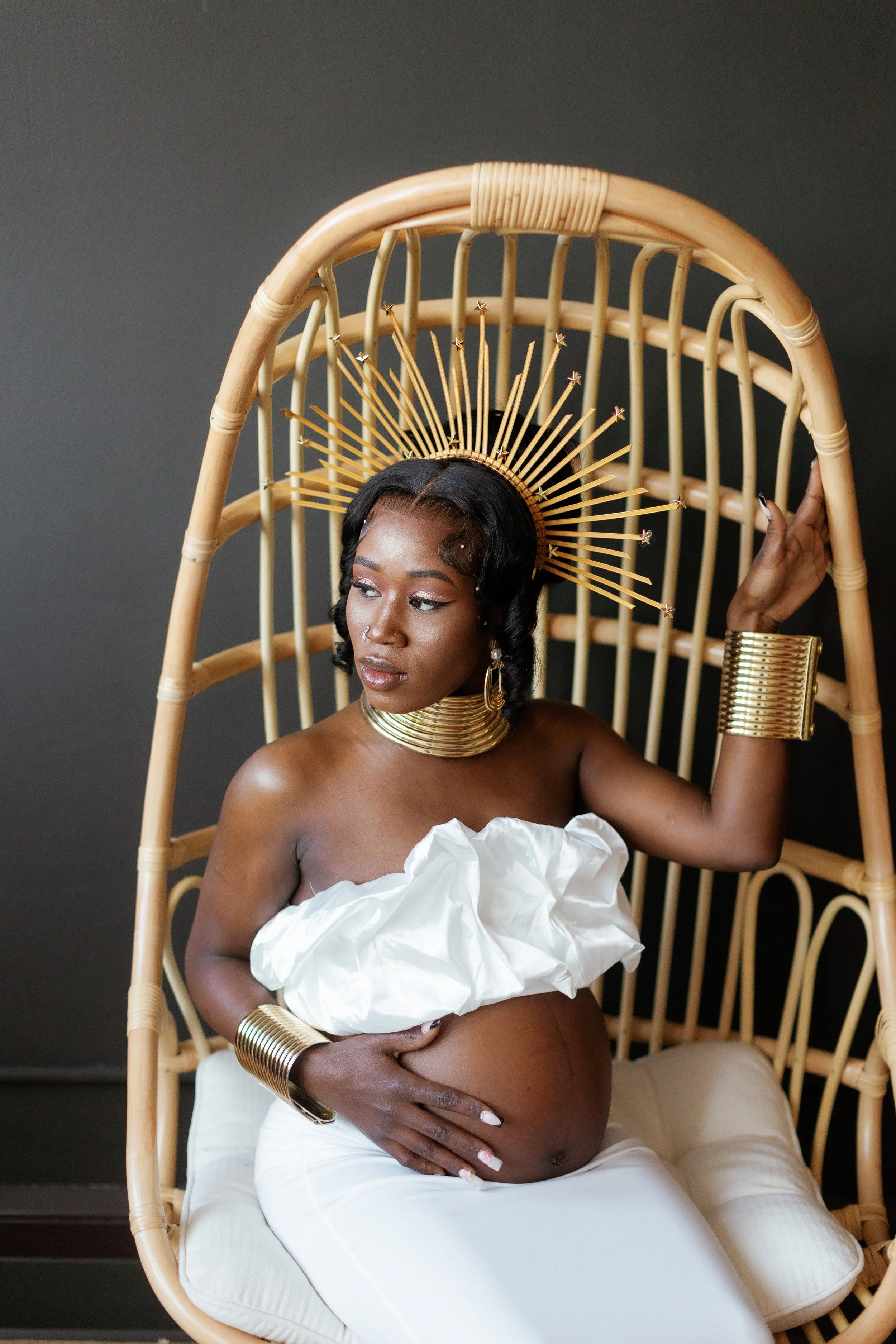 A woman with dark skin sitting in a wicker chair, wearing a white off-the-shoulder top and white pants. She has a large gold collar necklace, matching bracelets, and earrings. She is also wearing a decorative gold crown-like headpiece with radiating 