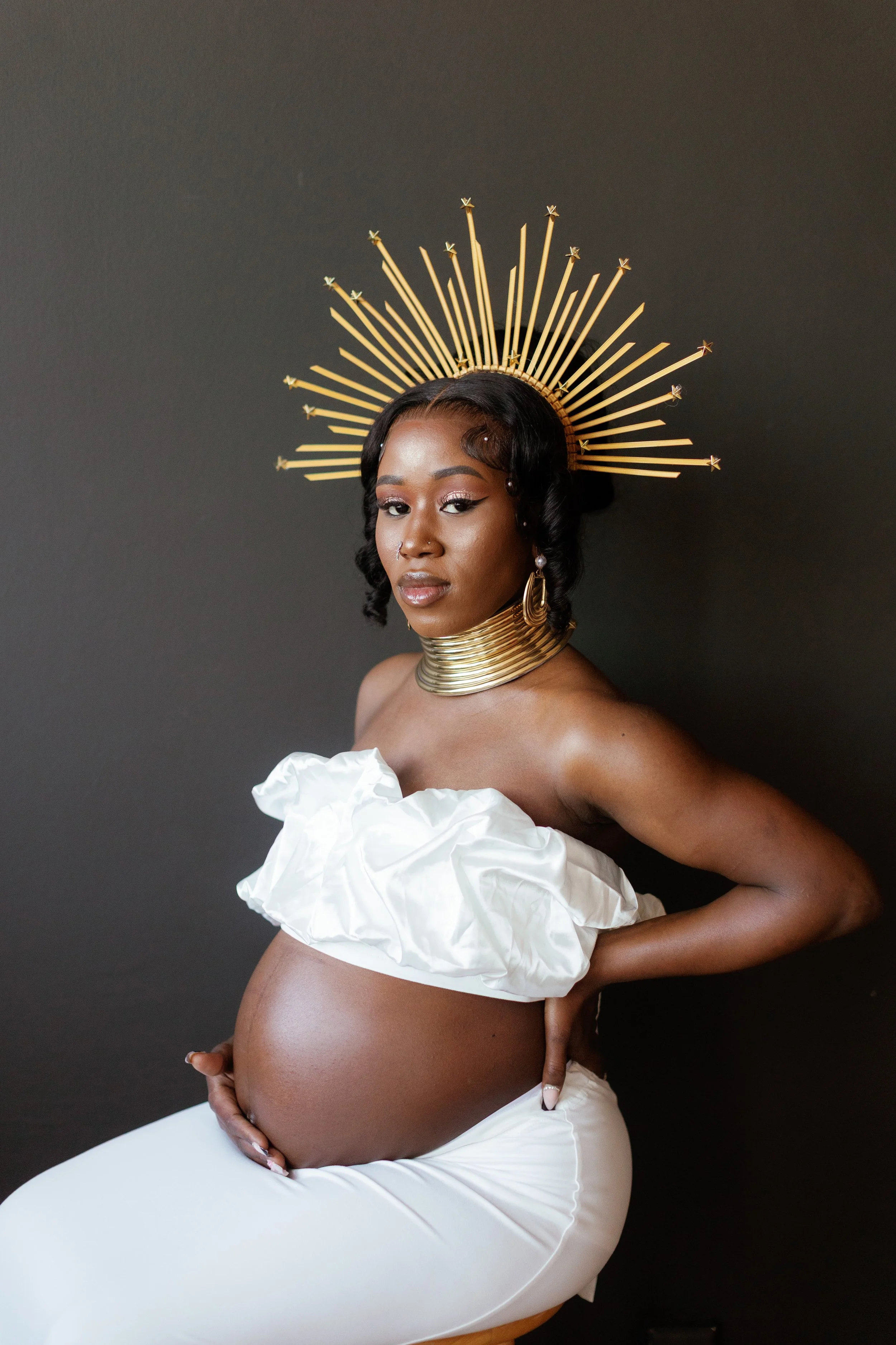 A pregnant woman with dark skin, wearing a white off-shoulder dress with ruffled fabric, is sitting against a plain dark background. She has styled hair with finger waves and wears gold jewelry including a wide collar necklace, dangly earrings, and a