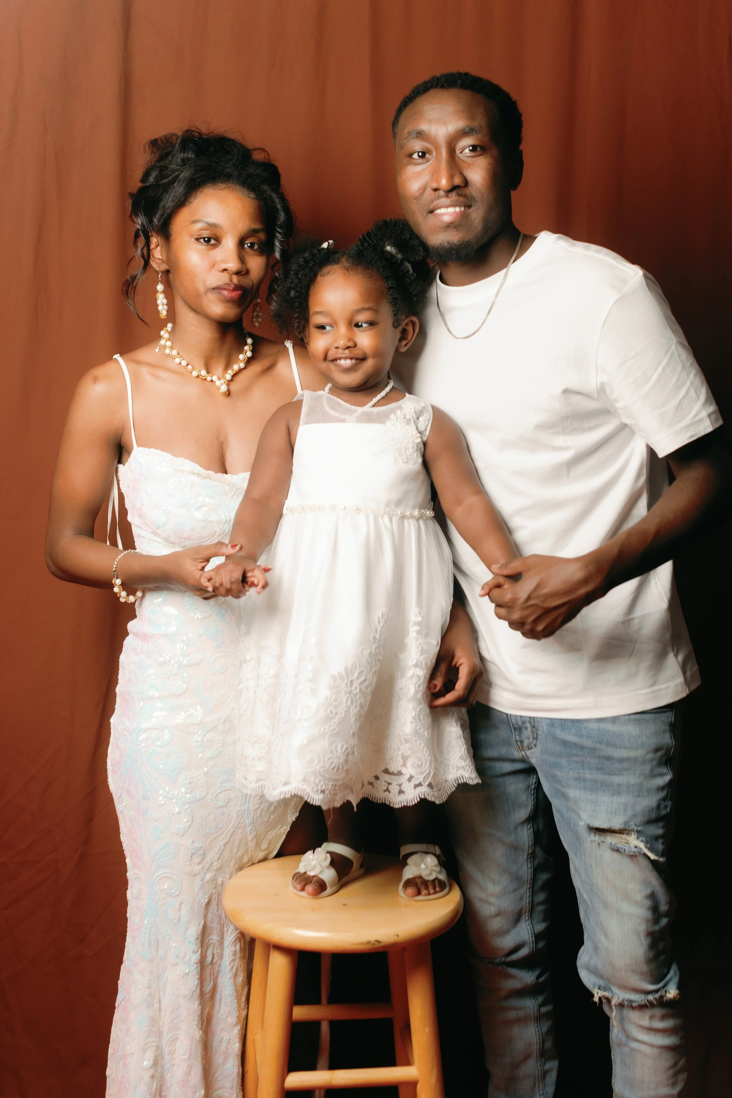 A family of three, a woman, a man, and a young girl, all dressed in white, standing together against a brown background. The woman is on the left, the man on the right, and the girl is standing on a wooden stool.