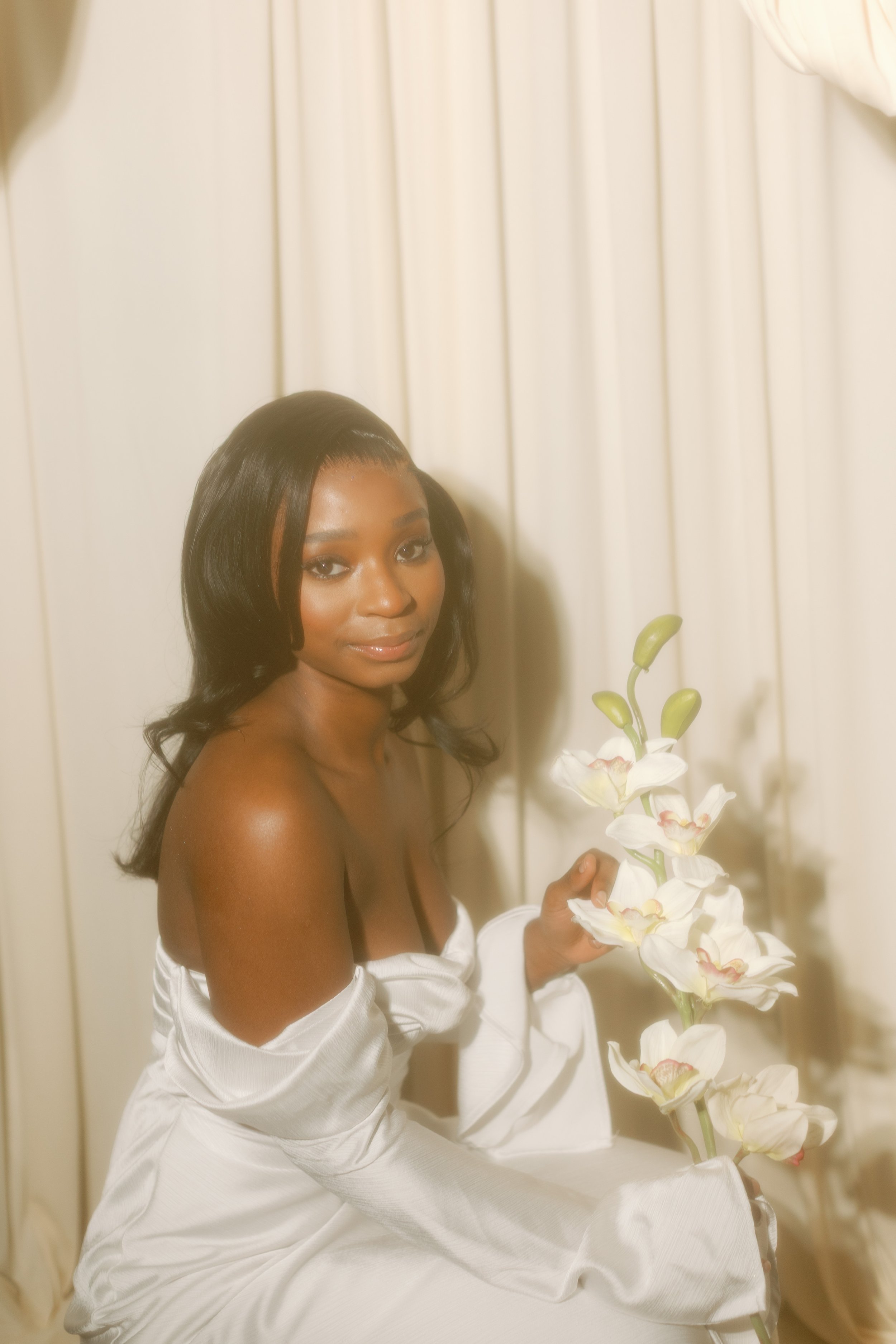 A woman with dark hair and dark skin, wrapped in a white towel, sitting against a cream-colored curtain, holding a tall white orchid plant with large white blossoms.