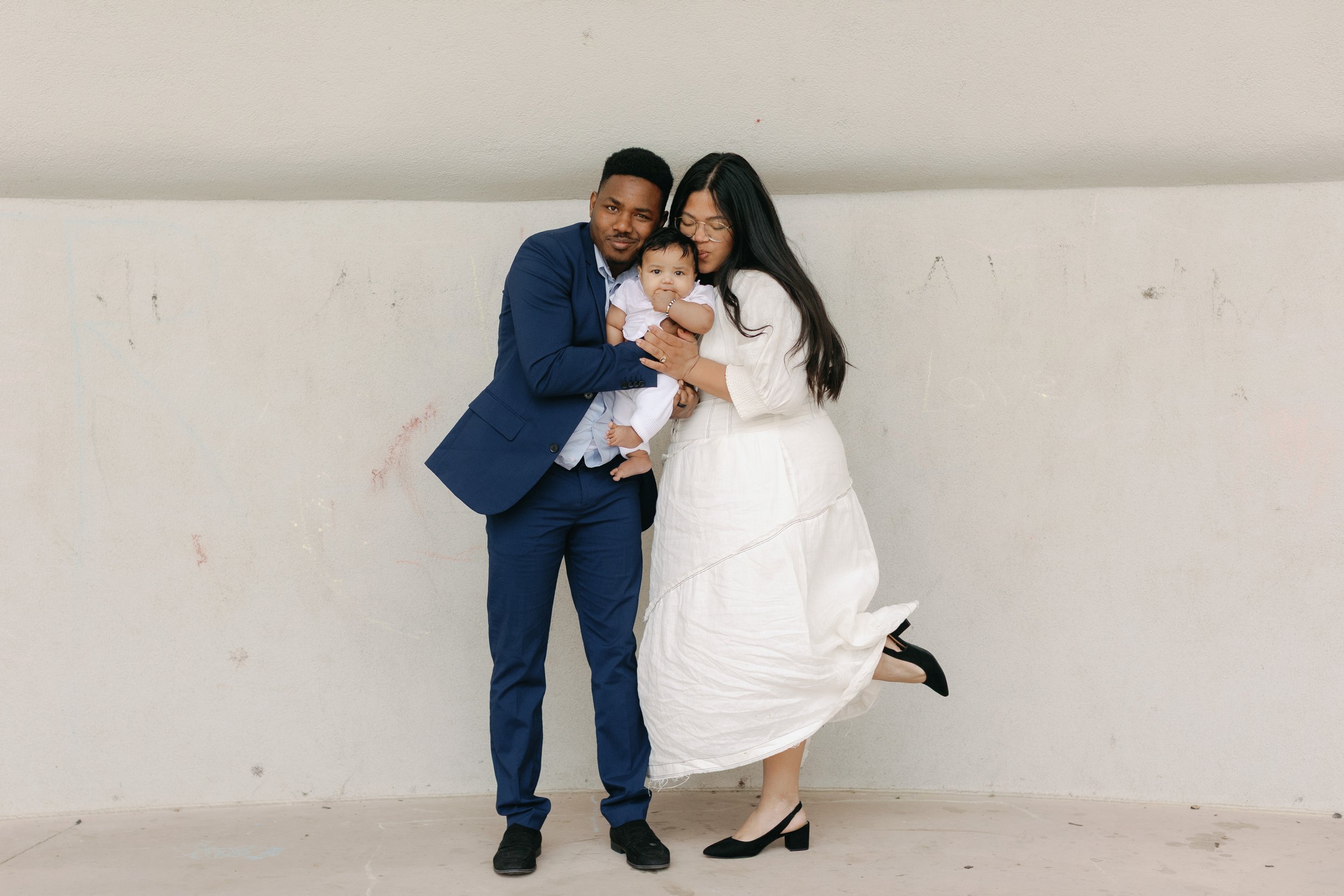 A family of three, including a man, a woman, and a baby, standing together against a plain wall. The man is wearing a blue suit, the woman is in a white dress, and the baby is dressed in white. The woman is holding the baby, and they are all smiling and close to each other.