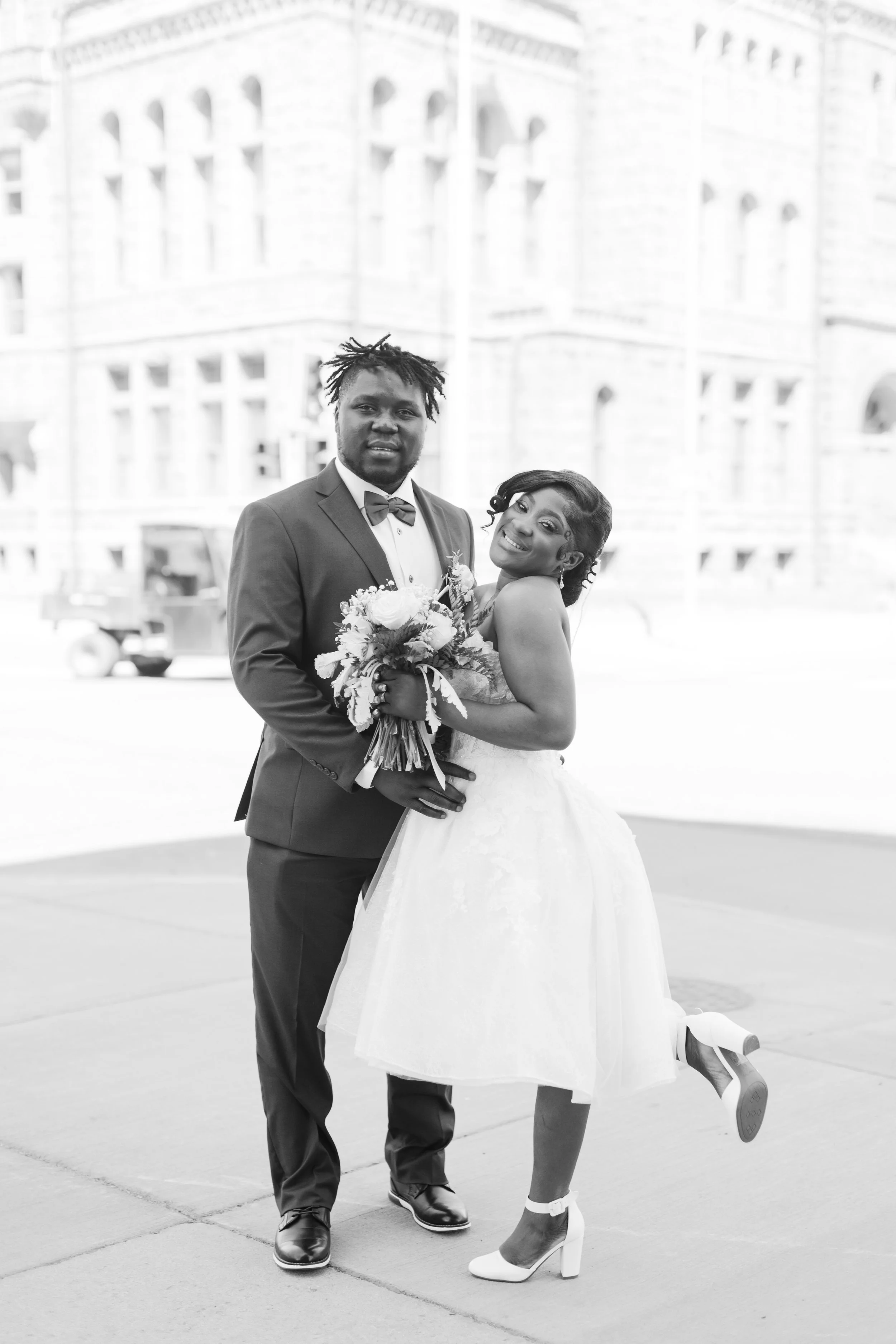 A black-and-white photo of a groom and bride posing together outdoors in front of a large building. The groom is wearing a suit with a bow tie, holding a bouquet, and the bride is in a strapless wedding dress, smiling and lifting one leg.