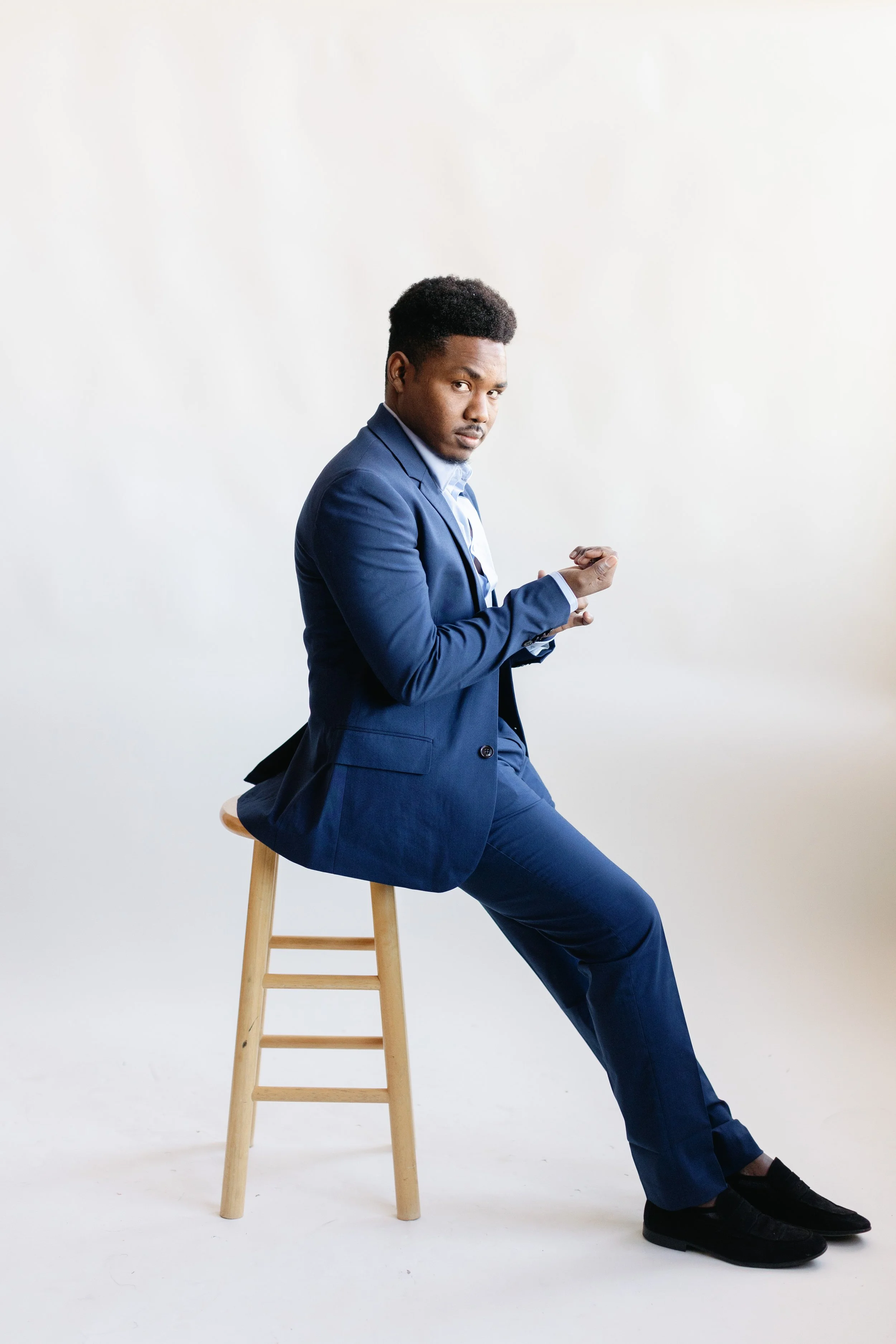 A man dressed in a blue suit sitting on a wooden stool against a plain white background, looking at the camera, with one leg crossed and arms resting on his lap.
