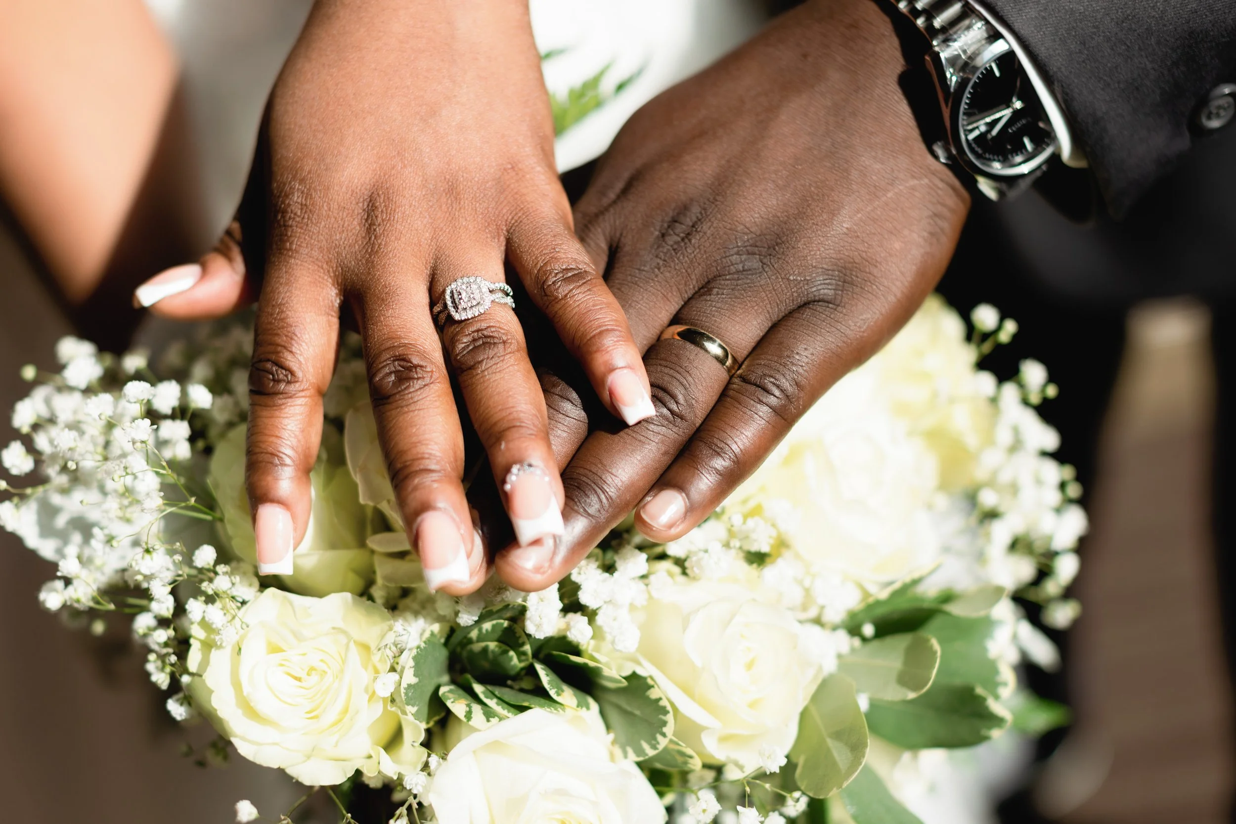 A close-up of two hands, one with darker skin and the other with lighter skin, resting together on a bouquet of white flowers. The darker-skinned hand is wearing a gold wedding band, and the lighter-skinned hand has a diamond engagement ring.