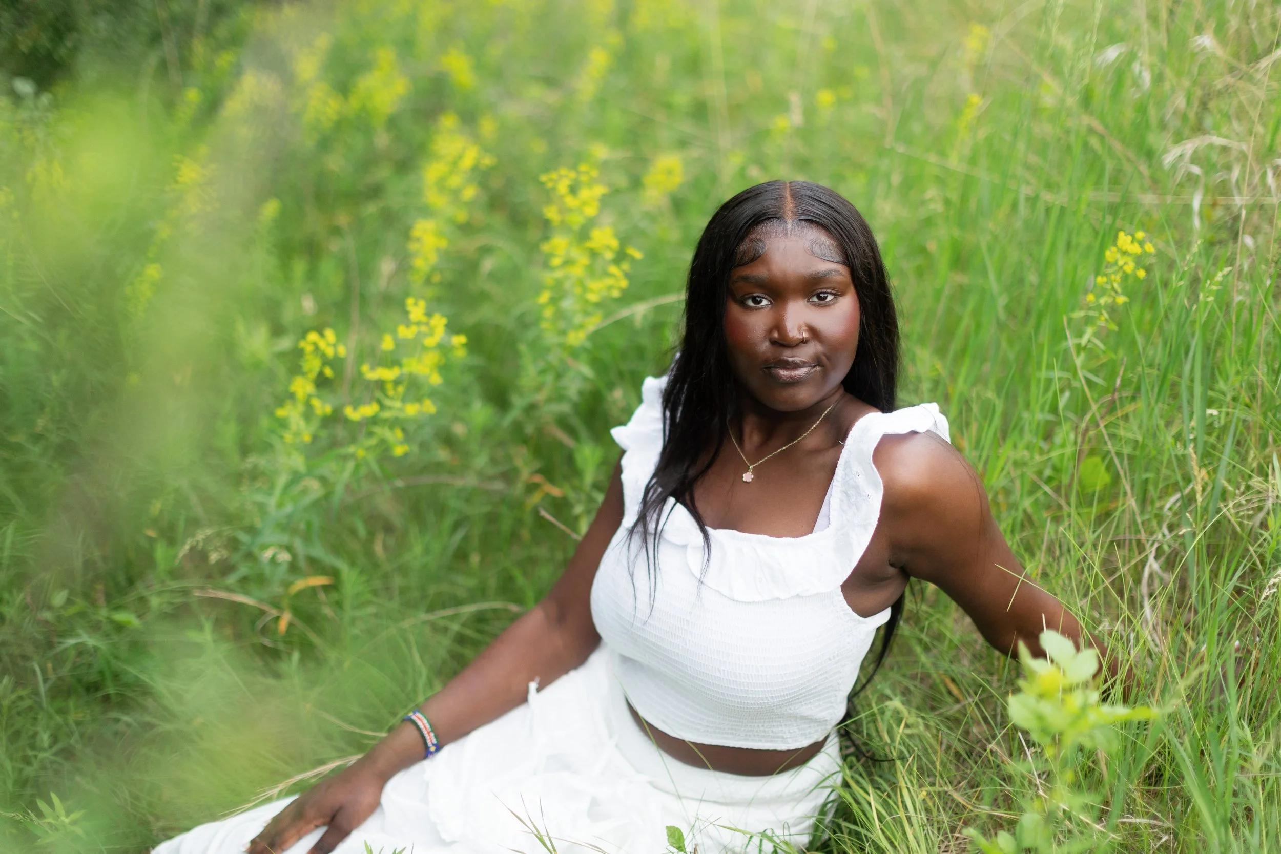 A young woman with dark hair and a nose piercing sitting in tall green grass outdoors, wearing a white sleeveless top and jewelry, surrounded by greenery.