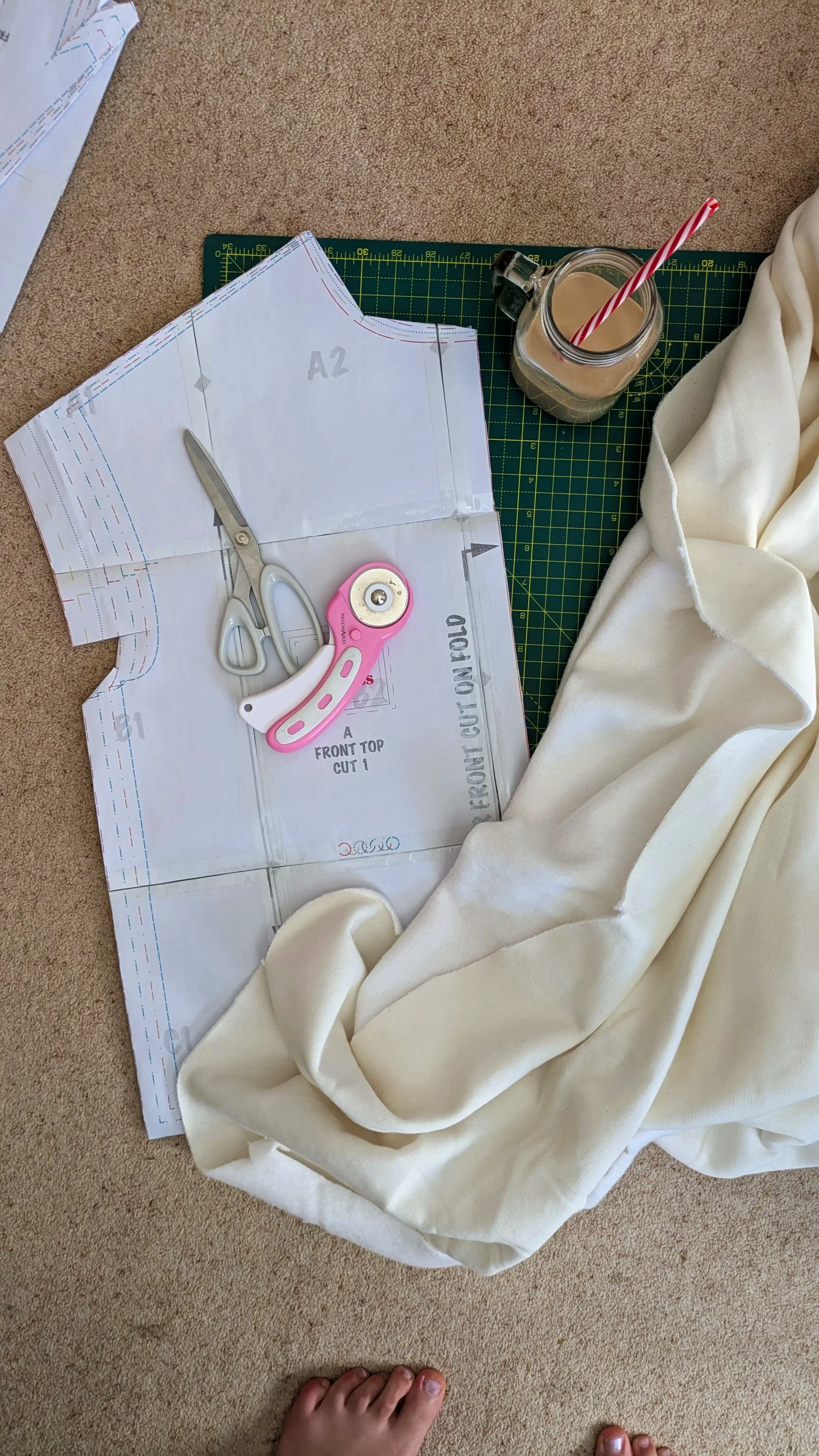 Sewing pattern paper on carpet with scissors, rotary cutter, fabric, mug with straw, and cutting mat.