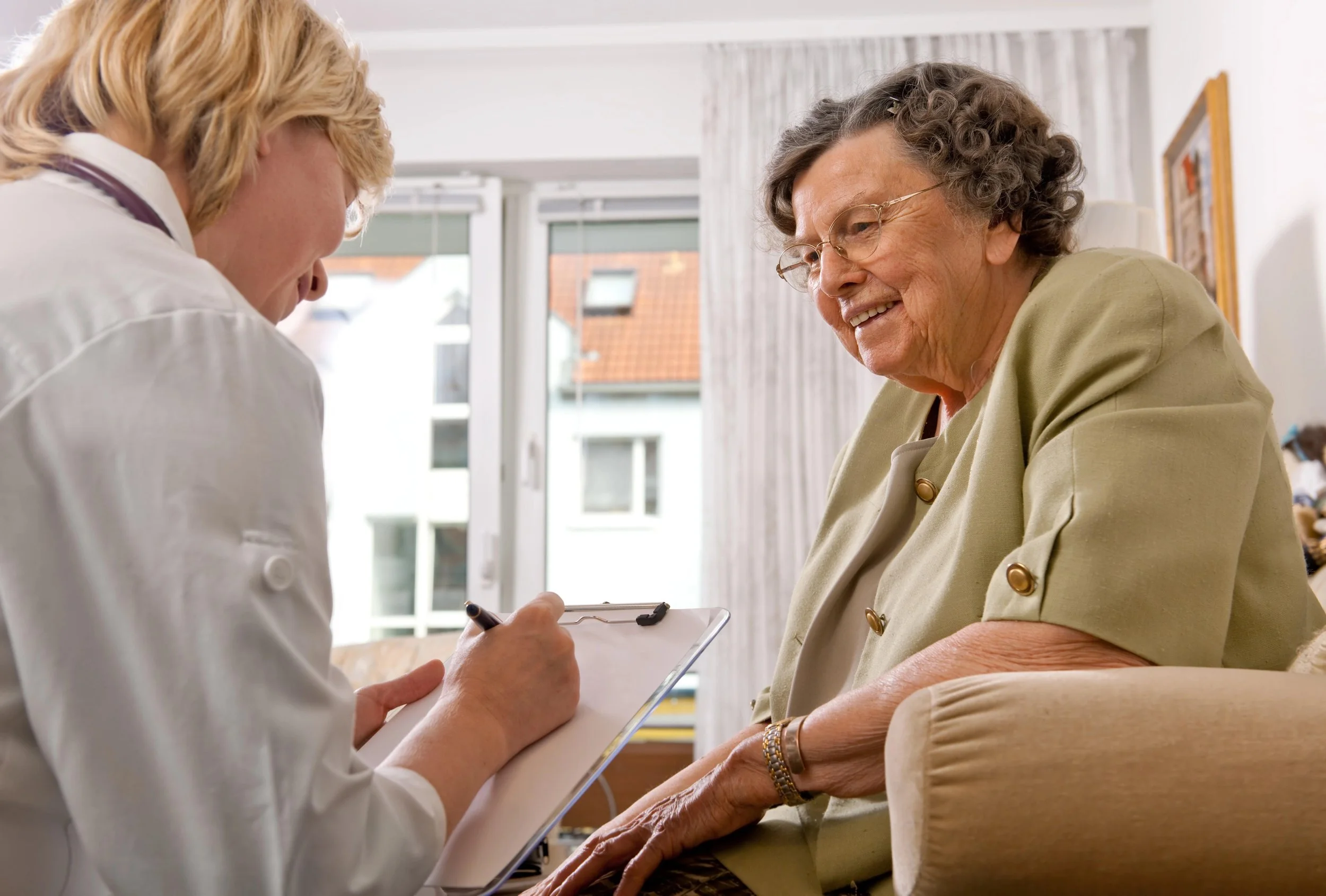 A nurse or healthcare worker writes on a clipboard while speaking to an elderly woman who is seated and smiling. The setting appears to be a healthcare facility or a home visit with large windows and natural light.