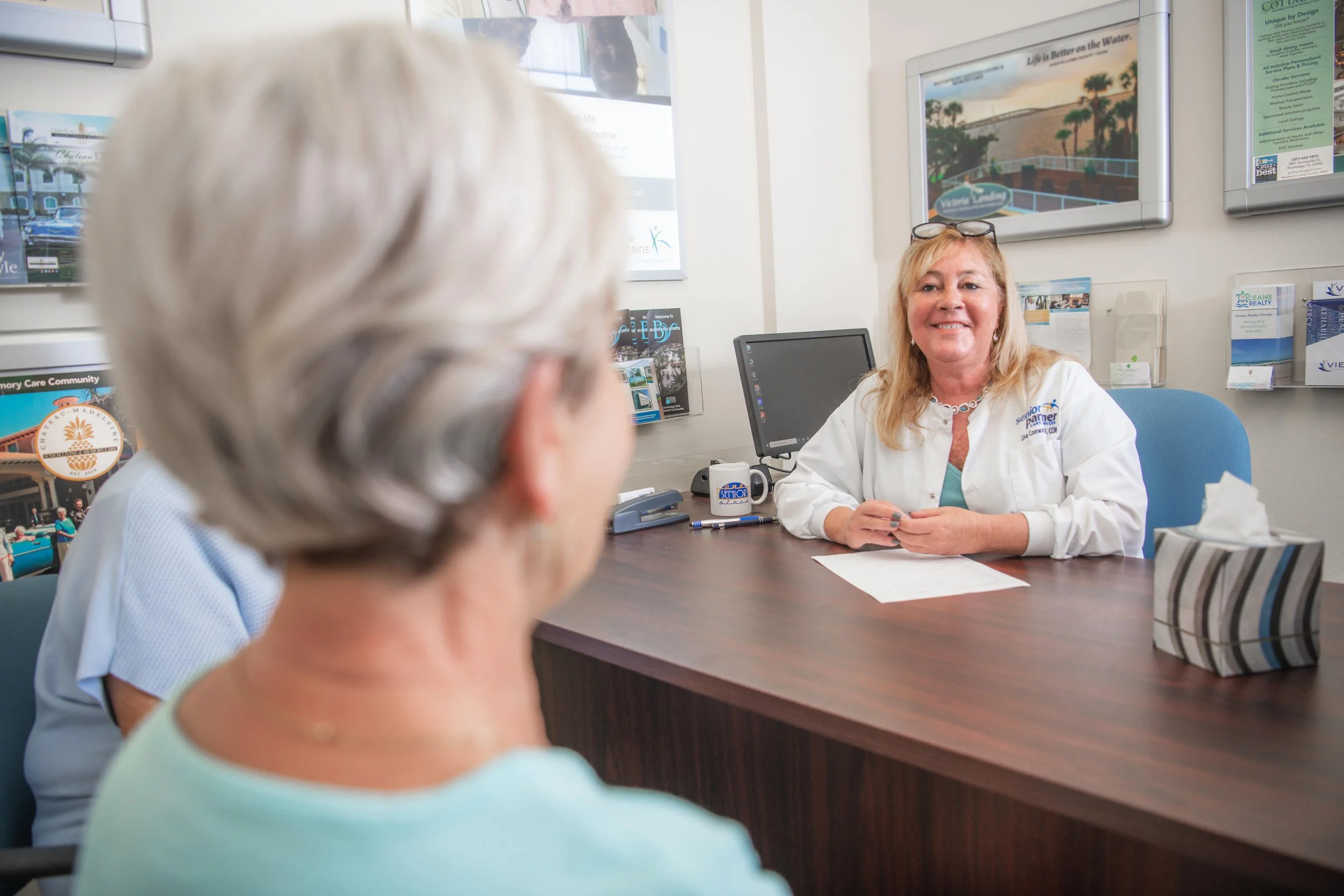 A woman with blonde hair, glasses on top of her head, and a white jacket sitting at a desk, smiling and talking to an elderly woman with gray hair in foreground, in an office setting with wall-mounted monitors, brochures, and a computer.