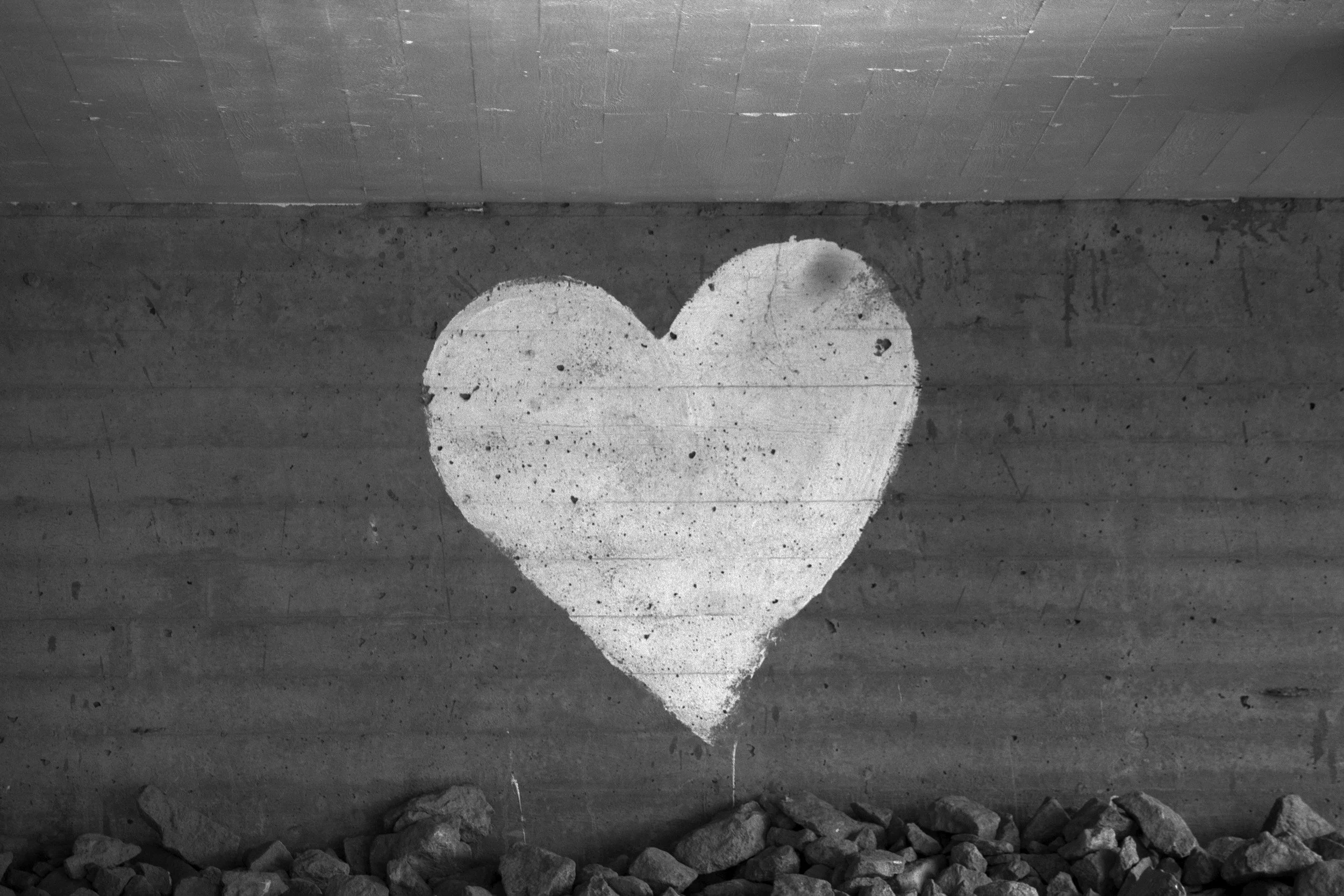 Black and white photograph of a concrete wall with a large painted white heart symbol, with rocks at the bottom of the wall.