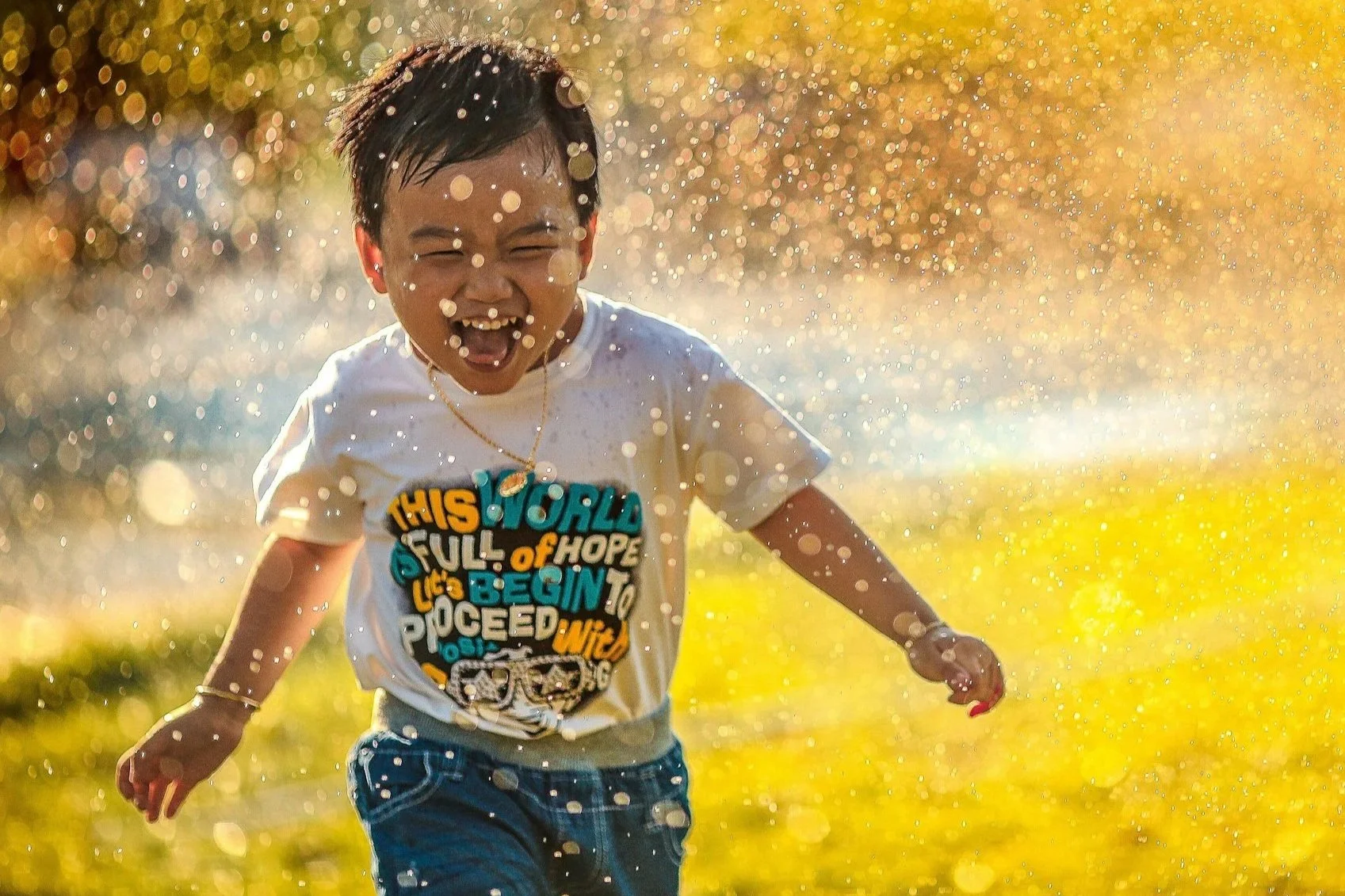 A young boy running joyfully through water in a field during golden hour, with sunlight illuminating the water droplets around him.