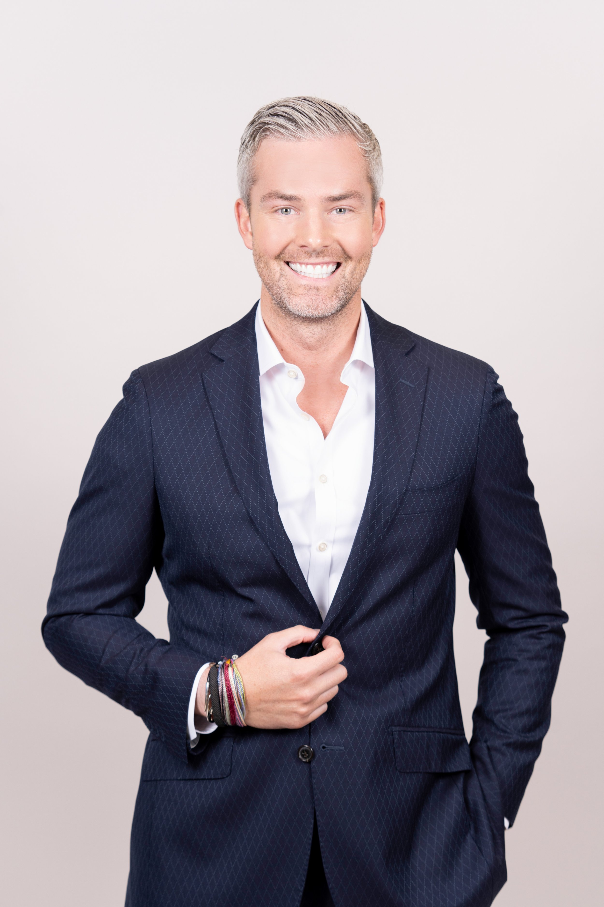 A man with light gray hair, wearing a navy blue patterned suit and white shirt, smiles at the camera against a plain light background.