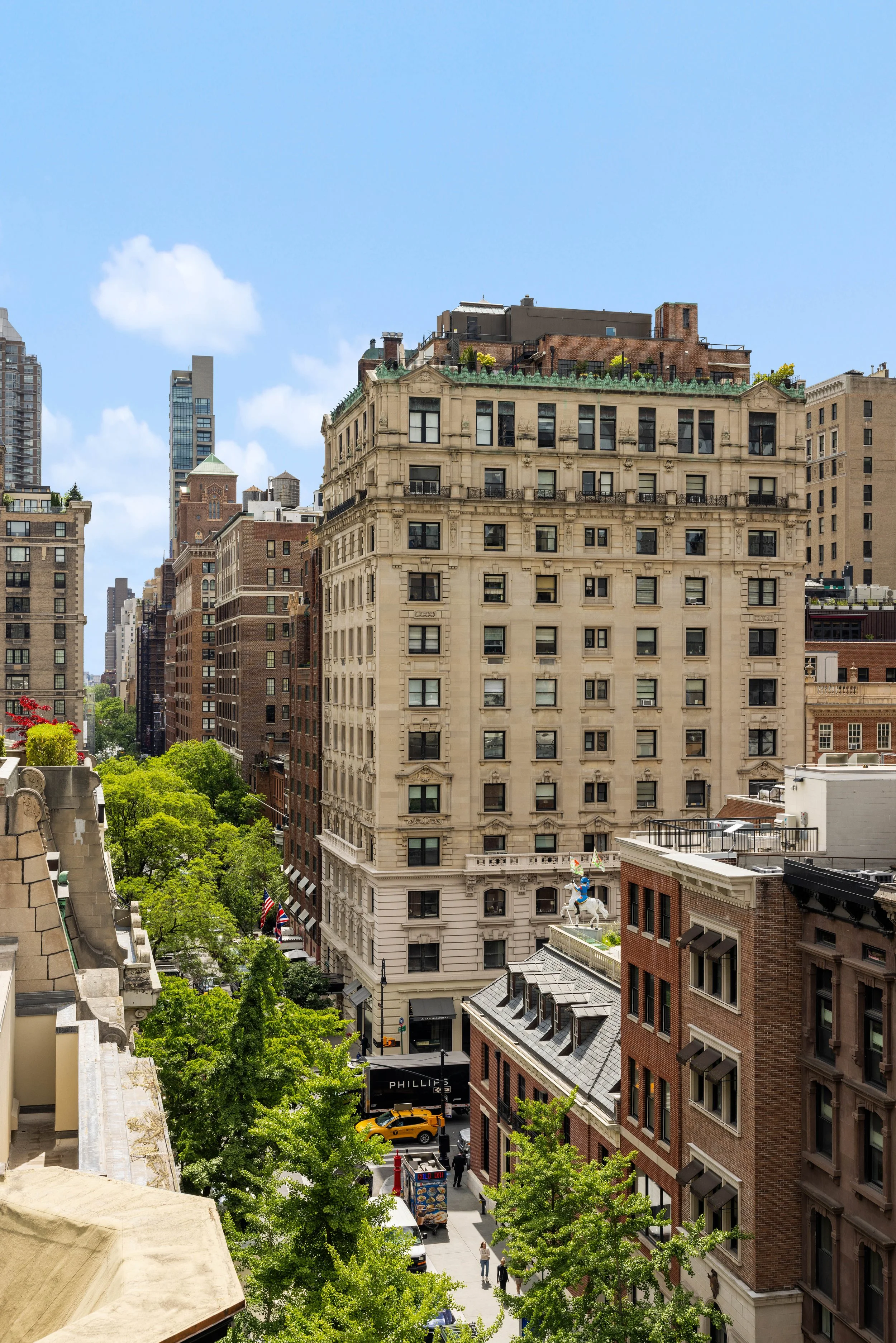 City street view with a mix of historic and modern buildings, trees lining the street, people walking, and vehicles including a yellow taxi and a food truck, under a blue sky with some clouds.