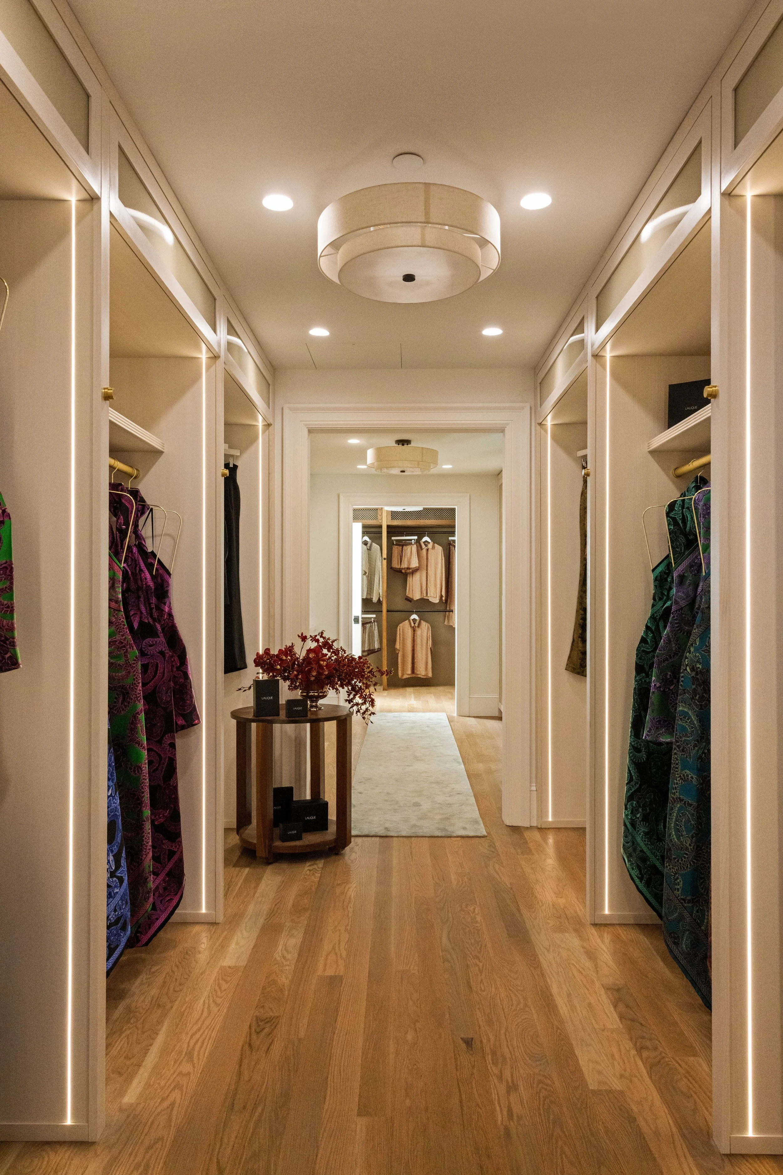 A hallway in a clothing store with hanging dresses on either side, a table with decoration in the middle, and a view of more clothing racks at the end.