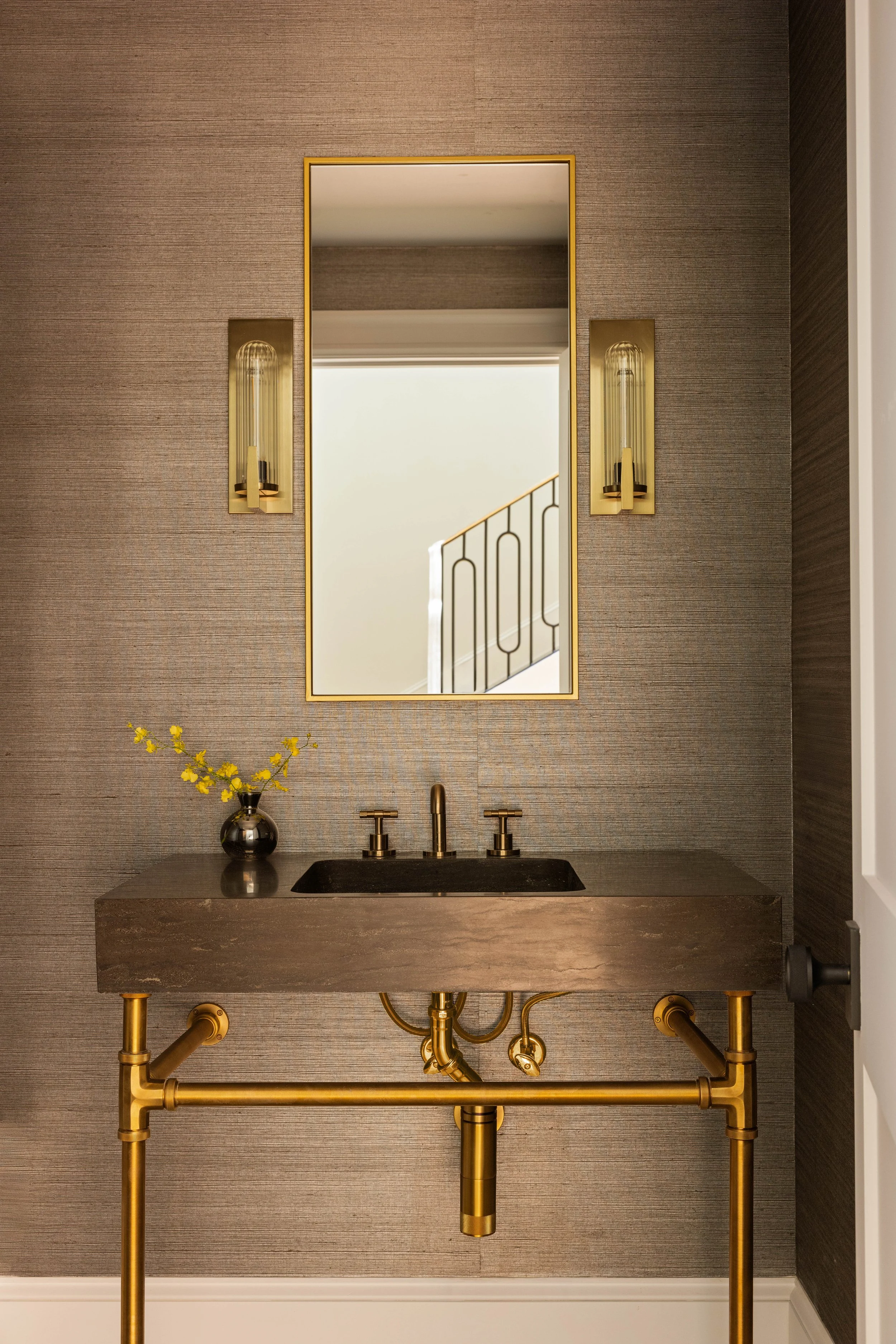 A modern bathroom vanity with a dark stone sink, gold faucet, and gold piping underneath. There is a mirror above the sink flanked by gold wall sconces, and a small bouquet of yellow flowers in a black vase on the left side of the vanity. The wall be