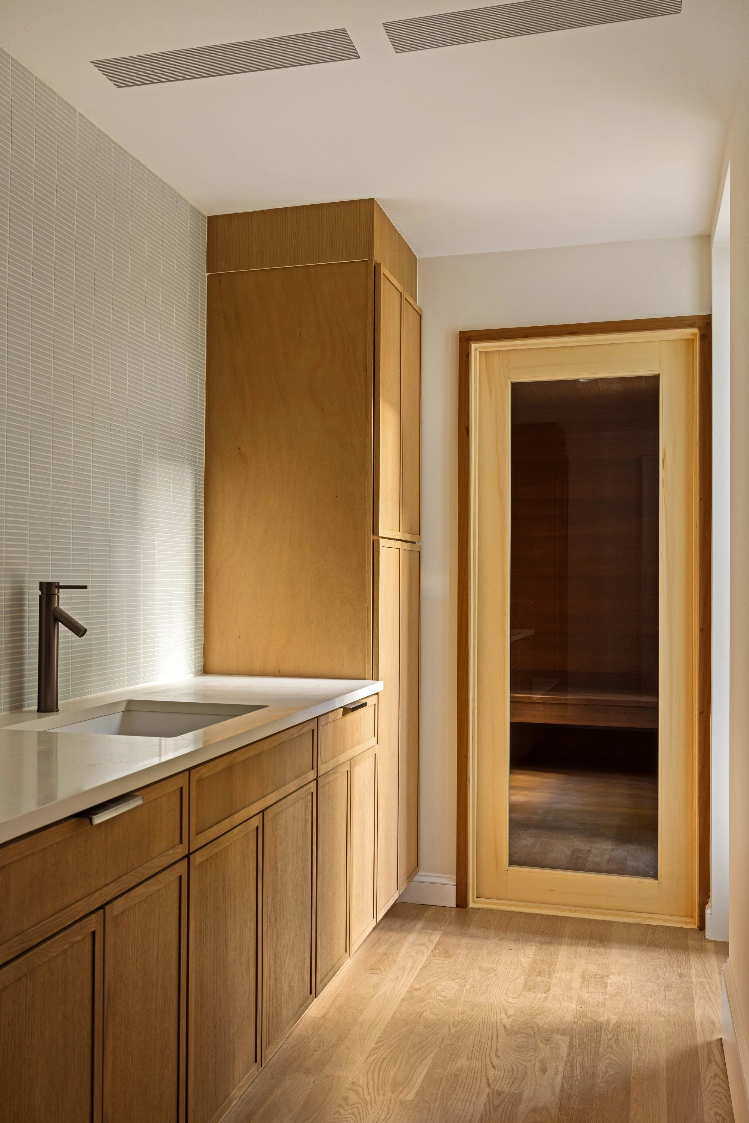 A kitchen with wooden cabinets, a white countertop, a sink with a black faucet, and a wooden framed glass door leading to another room with wooden floors.